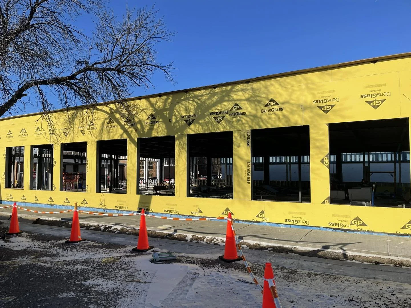 Building under construction with yellow insulation wrap, four large open window spaces, orange traffic cones, and a clear blue sky.