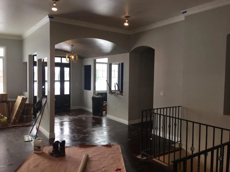 Living room and entryway under renovation with construction materials and tools visible, including cardboard boxes, boots, and a broom, with newly painted walls and windows.
