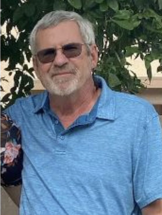 Older man with gray hair and beard wearing sunglasses and a blue polo shirt, standing outdoors with green foliage in the background.