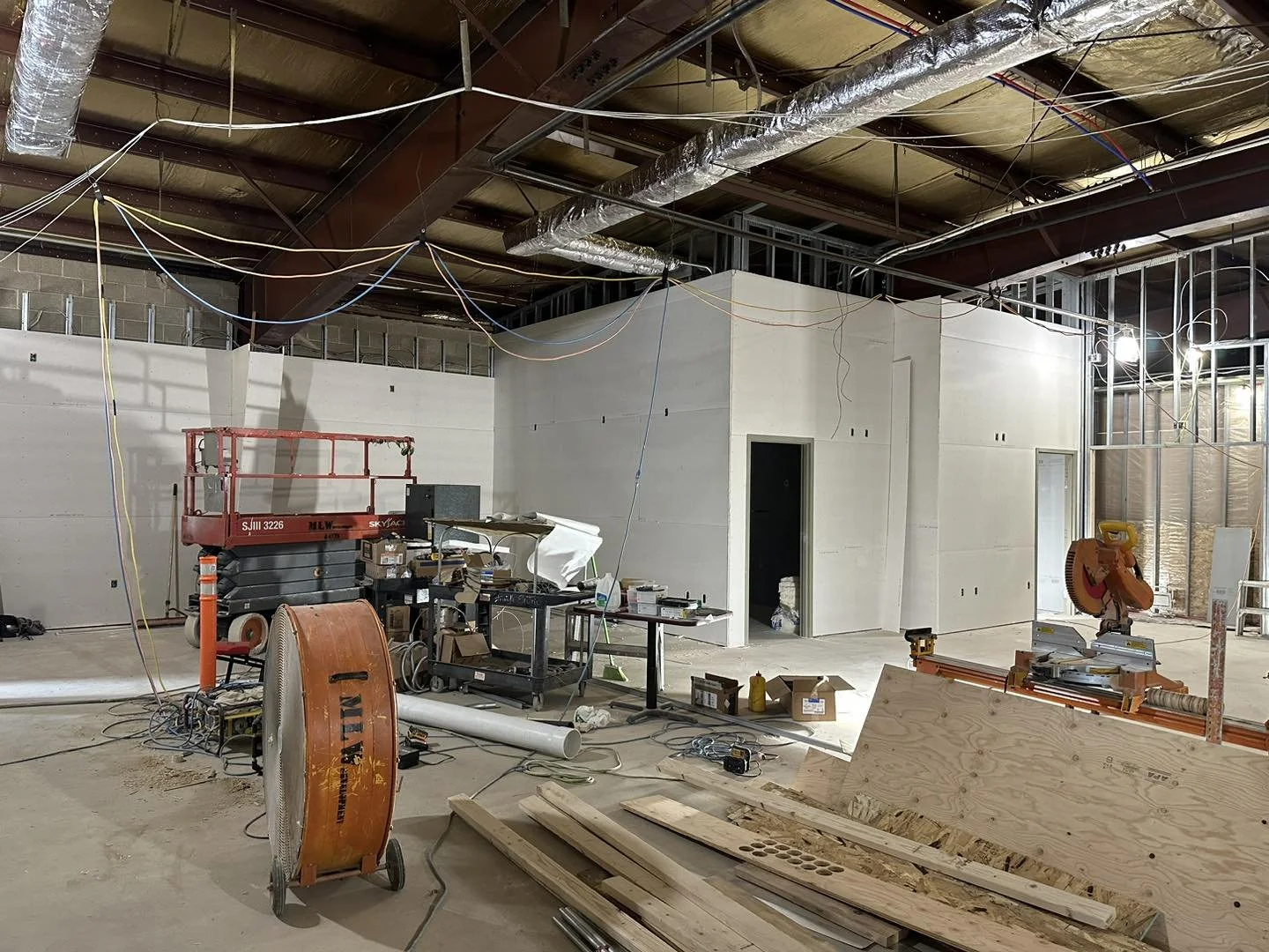 Construction site with unfinished walls, wiring hanging from the ceiling, and various tools and equipment scattered around.