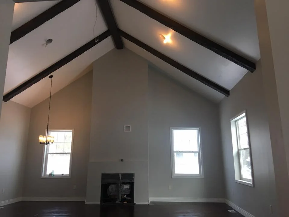 Interior of a living room with high vaulted ceiling, exposed dark wooden beams, three windows, and a small chandelier hanging from the ceiling.