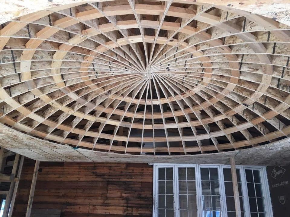 View of a circular wooden ceiling framework under construction, with radial and concentric beams, in a partially built interior room.