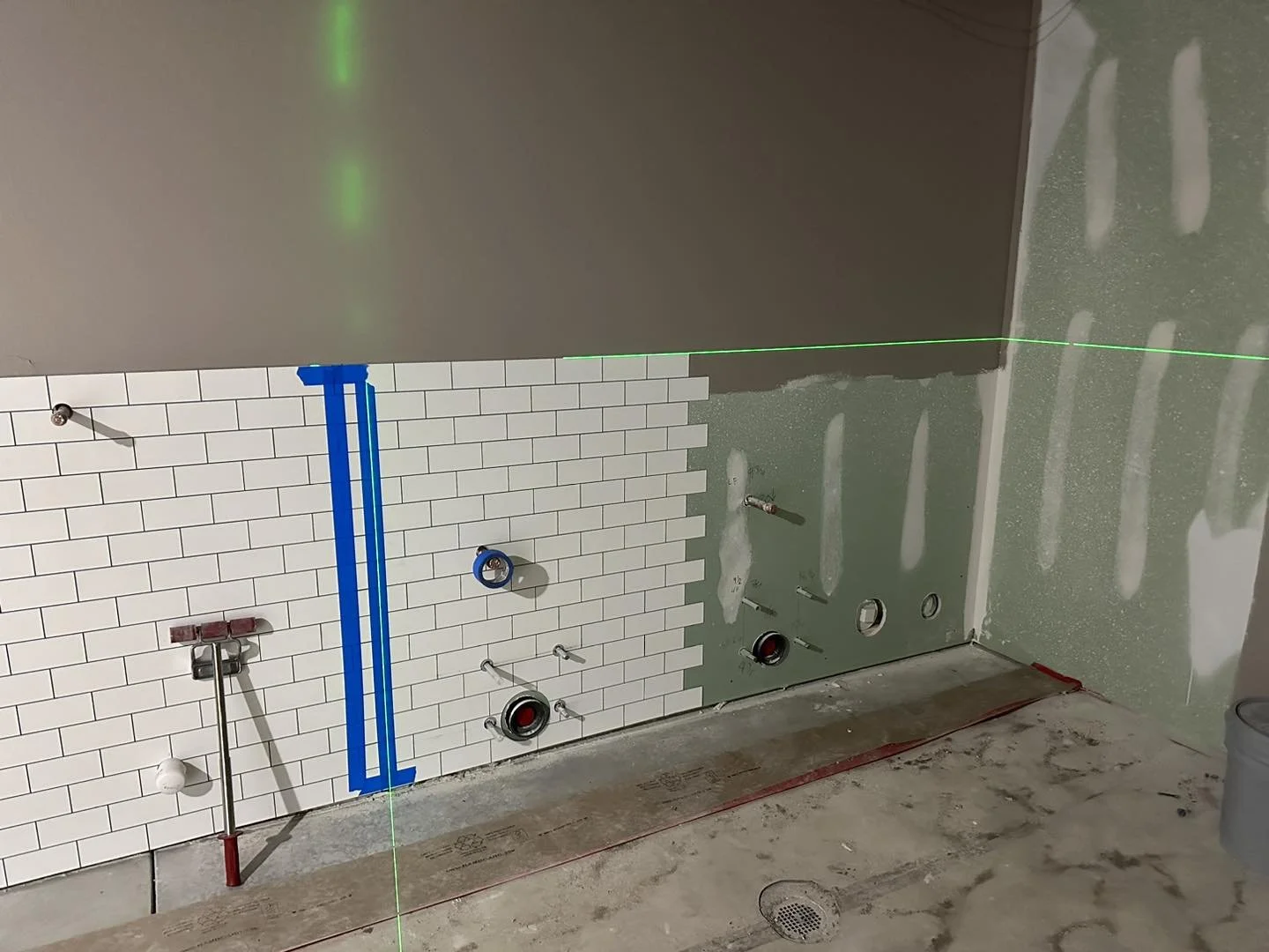 Bathroom under construction with partially tiled wall, exposed plumbing, and a laser level line across the unfinished space.
