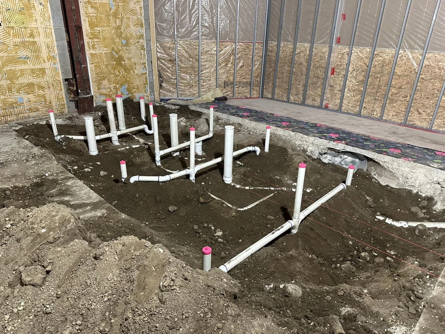 Construction site with exposed pipes and plumbing, dirt floor, and unfinished walls with insulation and wall sheathing.