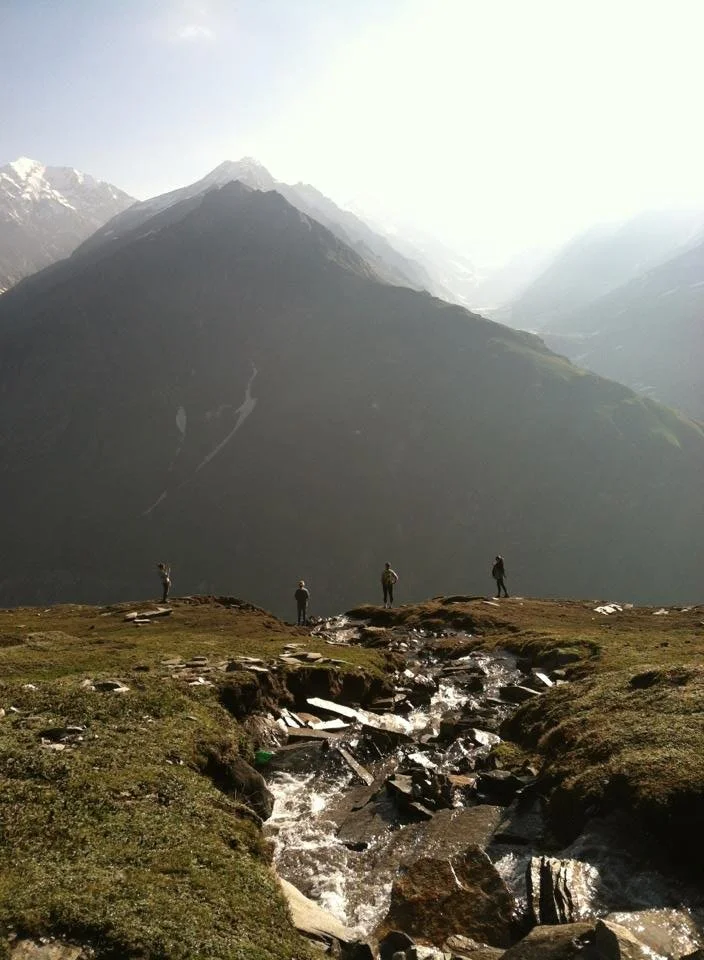a lush mountain scape with a rocky river in the foreground. four people stand looking at the mountain range with their backs to the camera.
