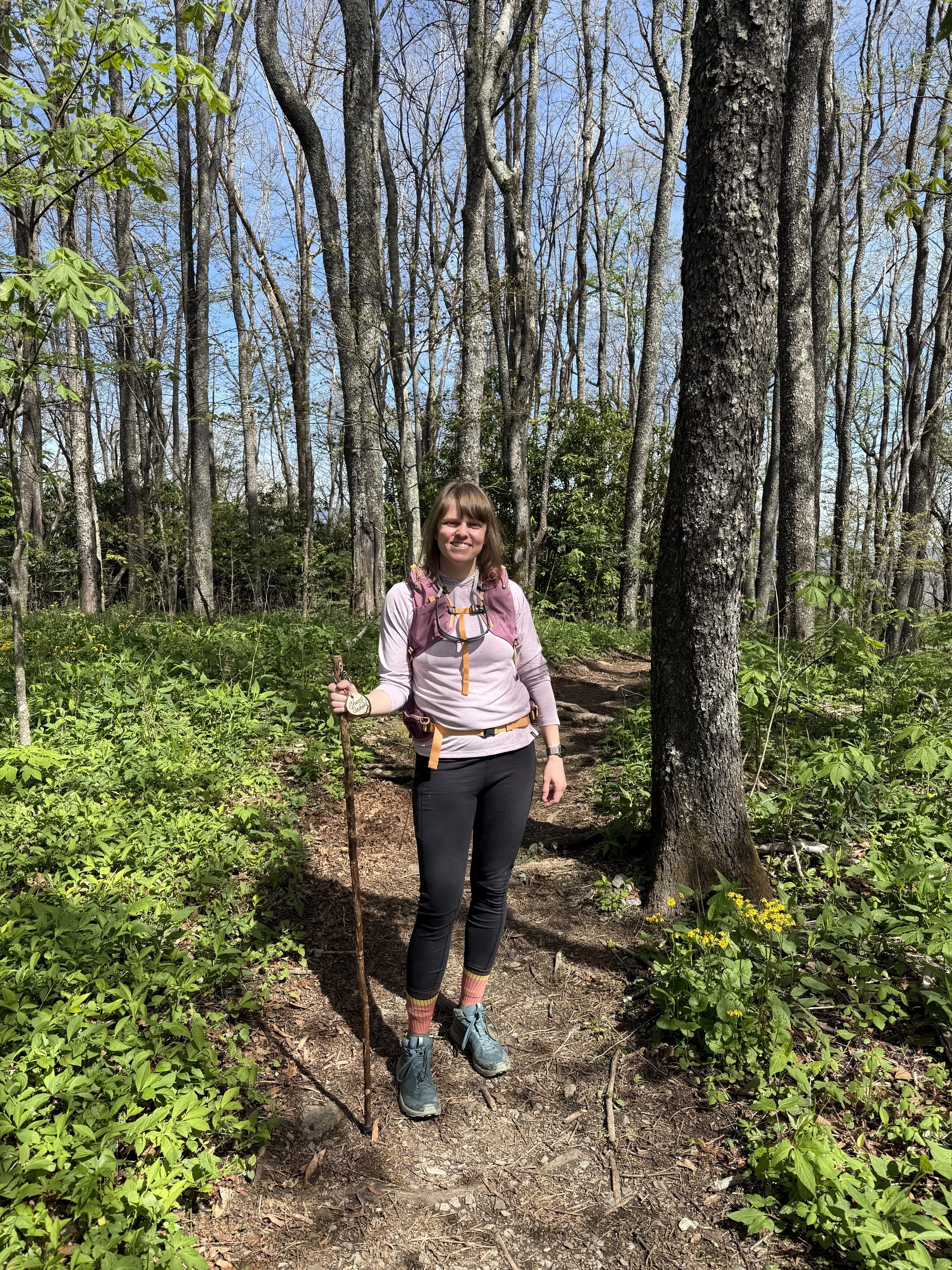 a whit woman with medium brown hair dressed in hiking gear holds a walking stick and poses for a picture in the woods. It is early spring so there is some greenery but the trees are not in bloom yet.