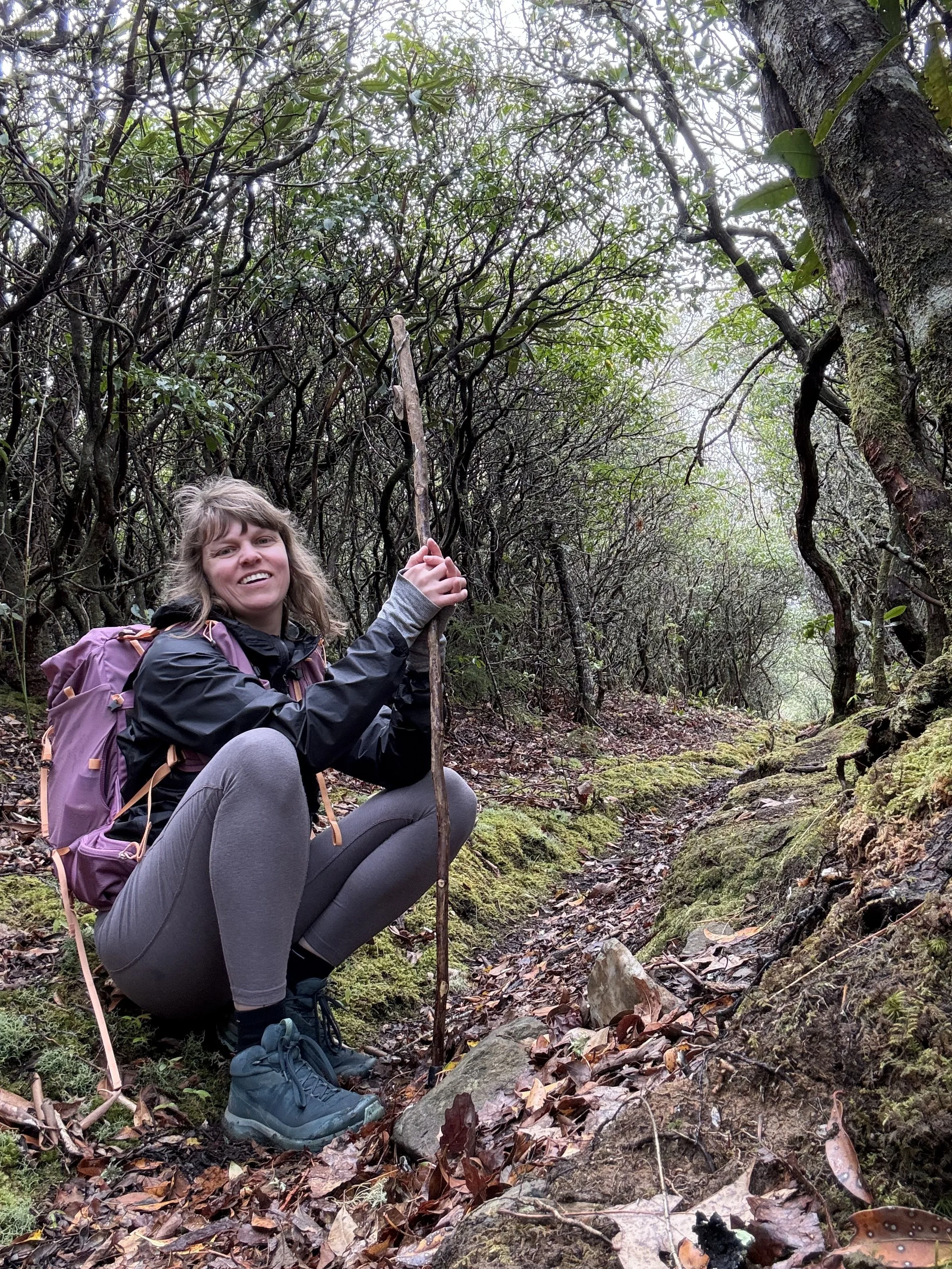 a middle aged white woman with bangs and medium brown hair in hiking gear is crouched holding a walking stick and smiles for the camera. The backdrop is lush with green moss covered rocks and trees.
