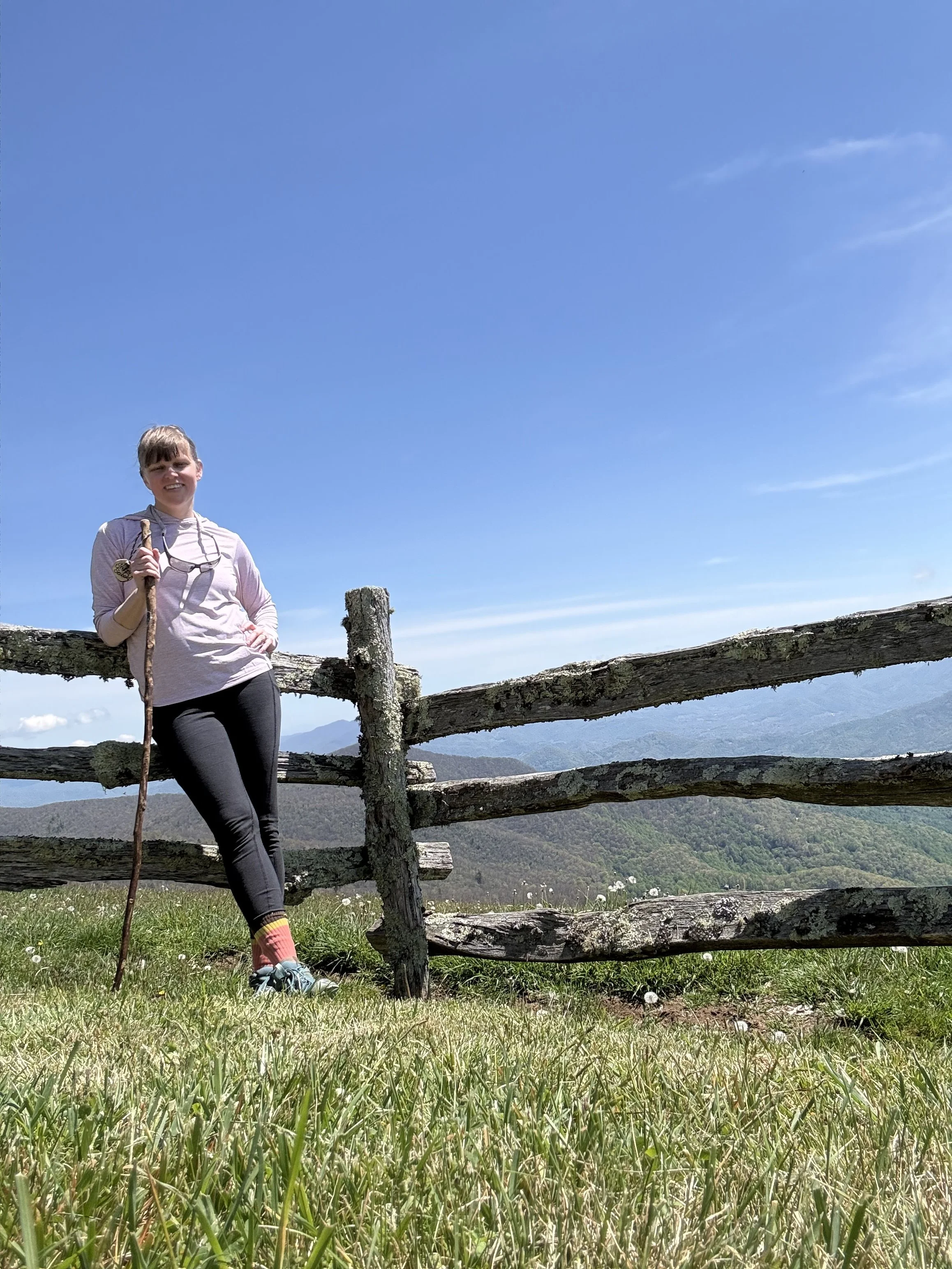 a woman posing in front on a fence with a mountainrange in the background. She has completed a hike and smiles at the camera. She is wearing a purple top and black pants.