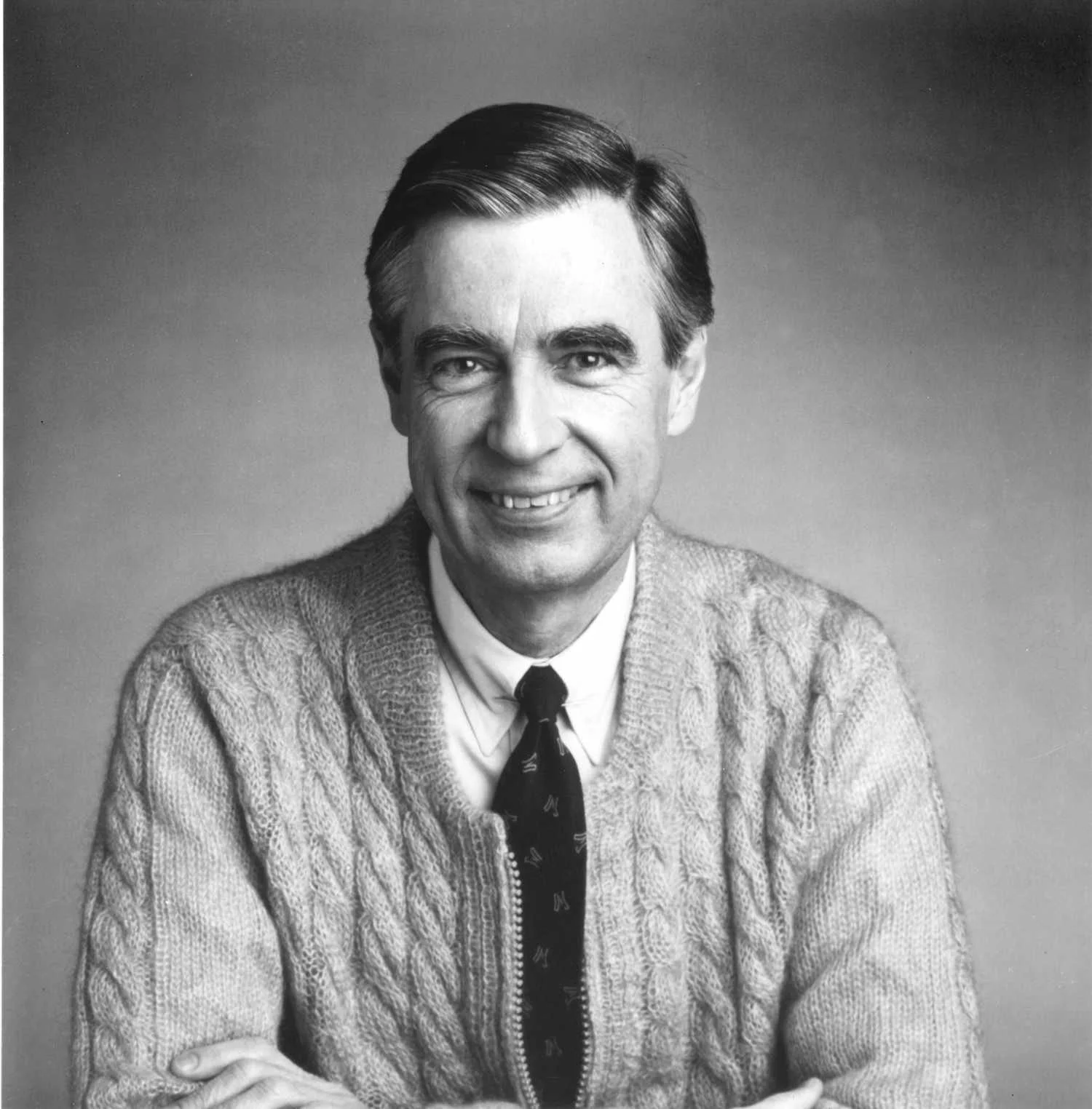 A black and white photo of Mr. Rogers- a white man wearing a sweater over a button down shirt and tie. He is smiling at the camera.