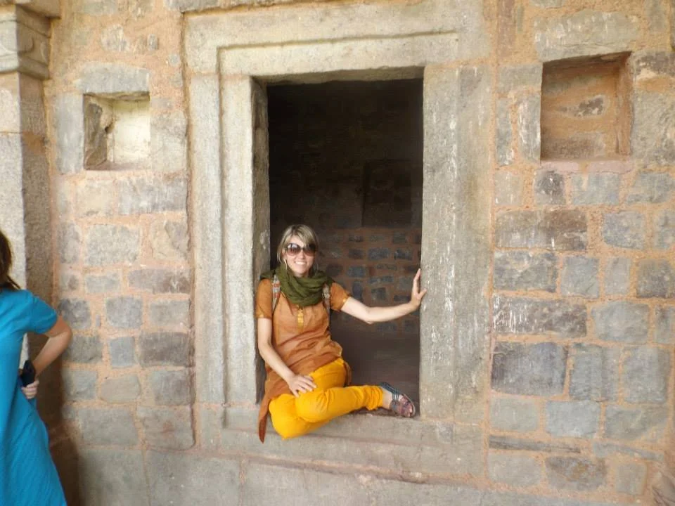a young adult woman wearing yellow pants, a golden orange tunic, olive green scarf, and sunglasses poses in a window sill of an old stone building in India.