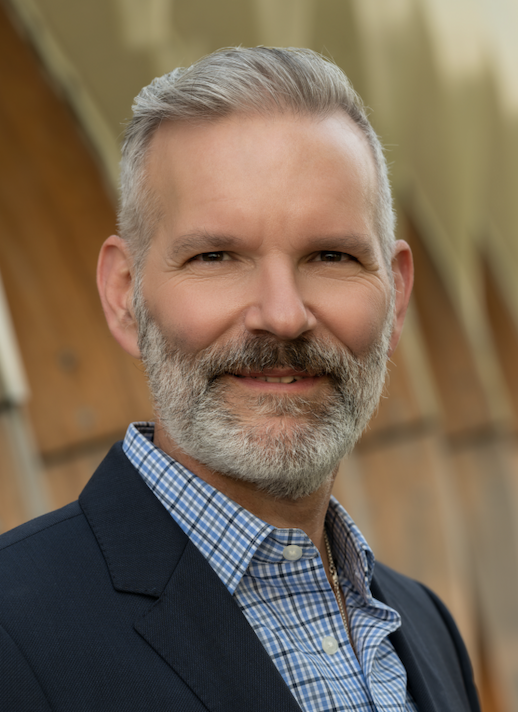 a man with a beard wearing a blue button down shirt and jacket smiles at the camera