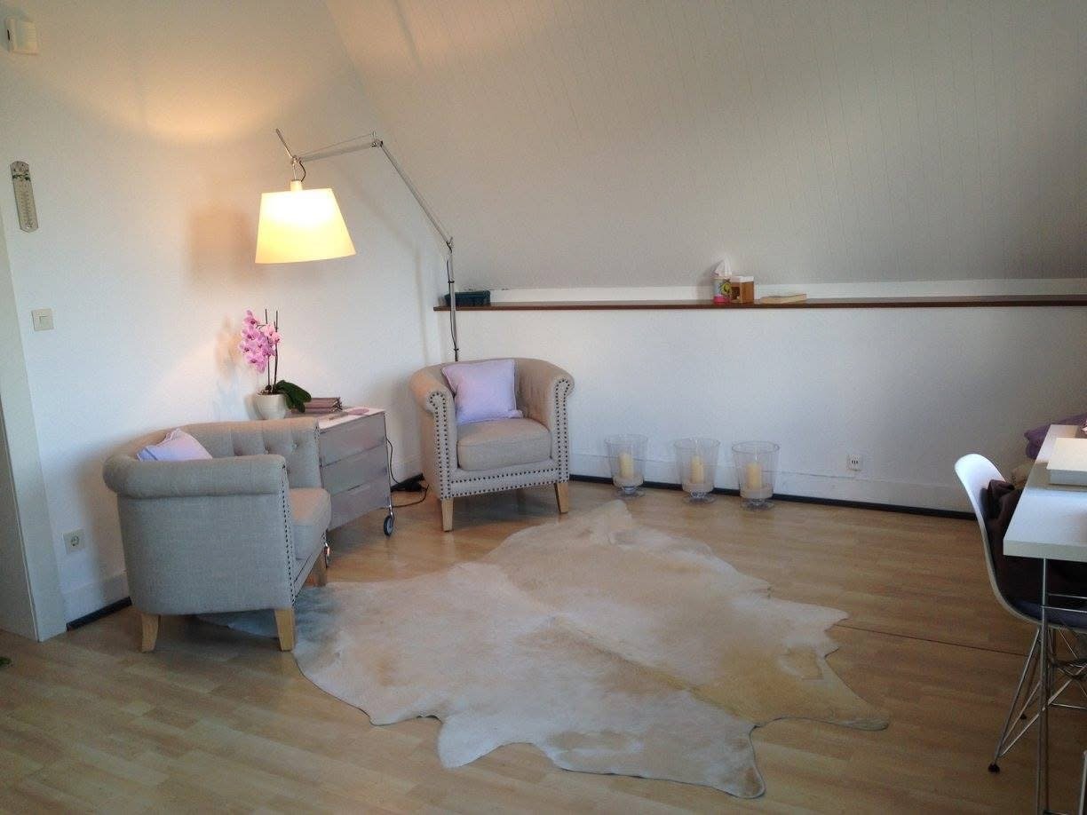 Gemütliches Wohnzimmer mit zwei beige Sessel, einem weißen Teppich, einem Beistelltisch mit einer Orchidee, einer Stehlampe, Kerzen auf dem Boden und einer weißen Wand.