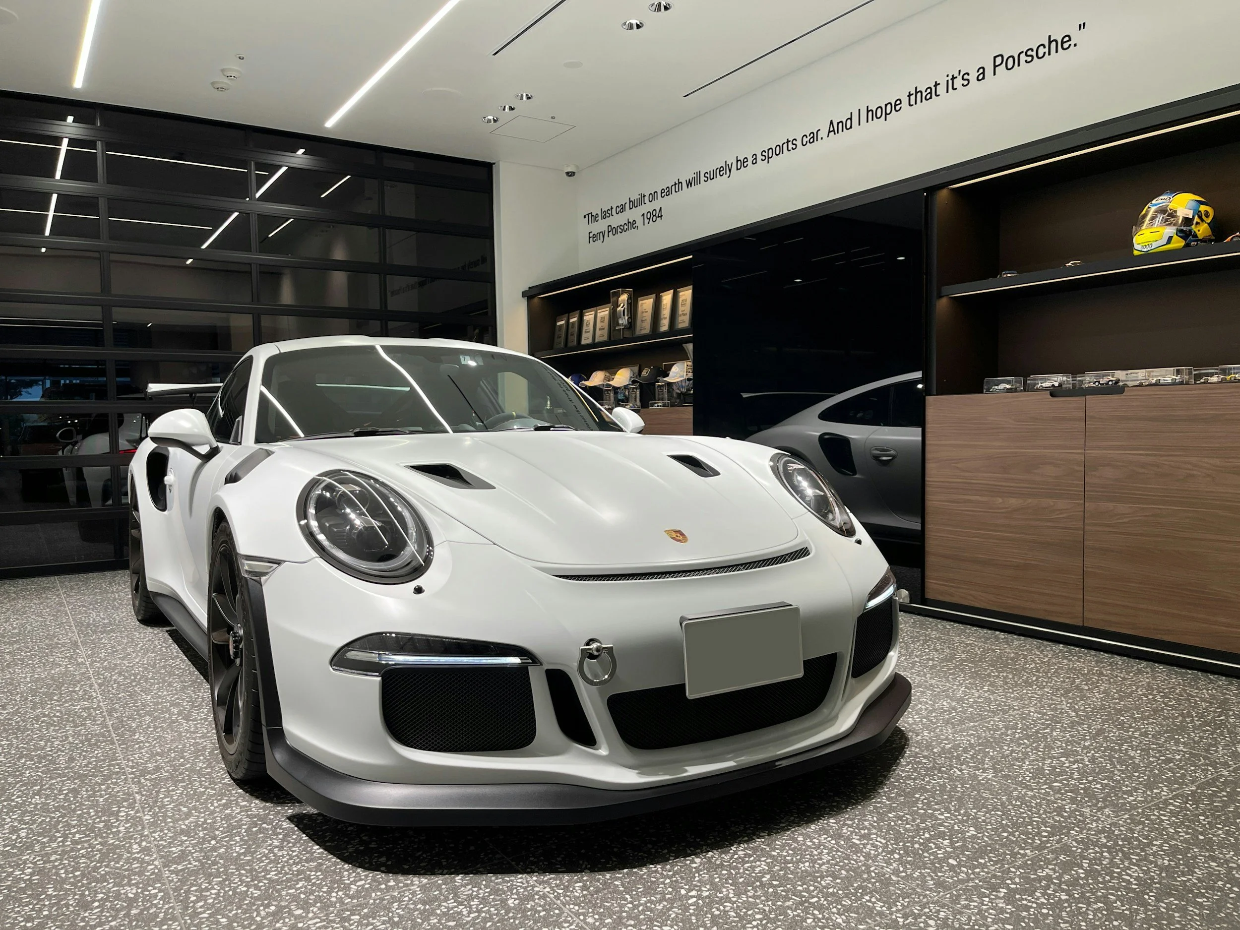 A white Porsche sports car on display inside a modern showroom with quotes about Porsche on the wall.
