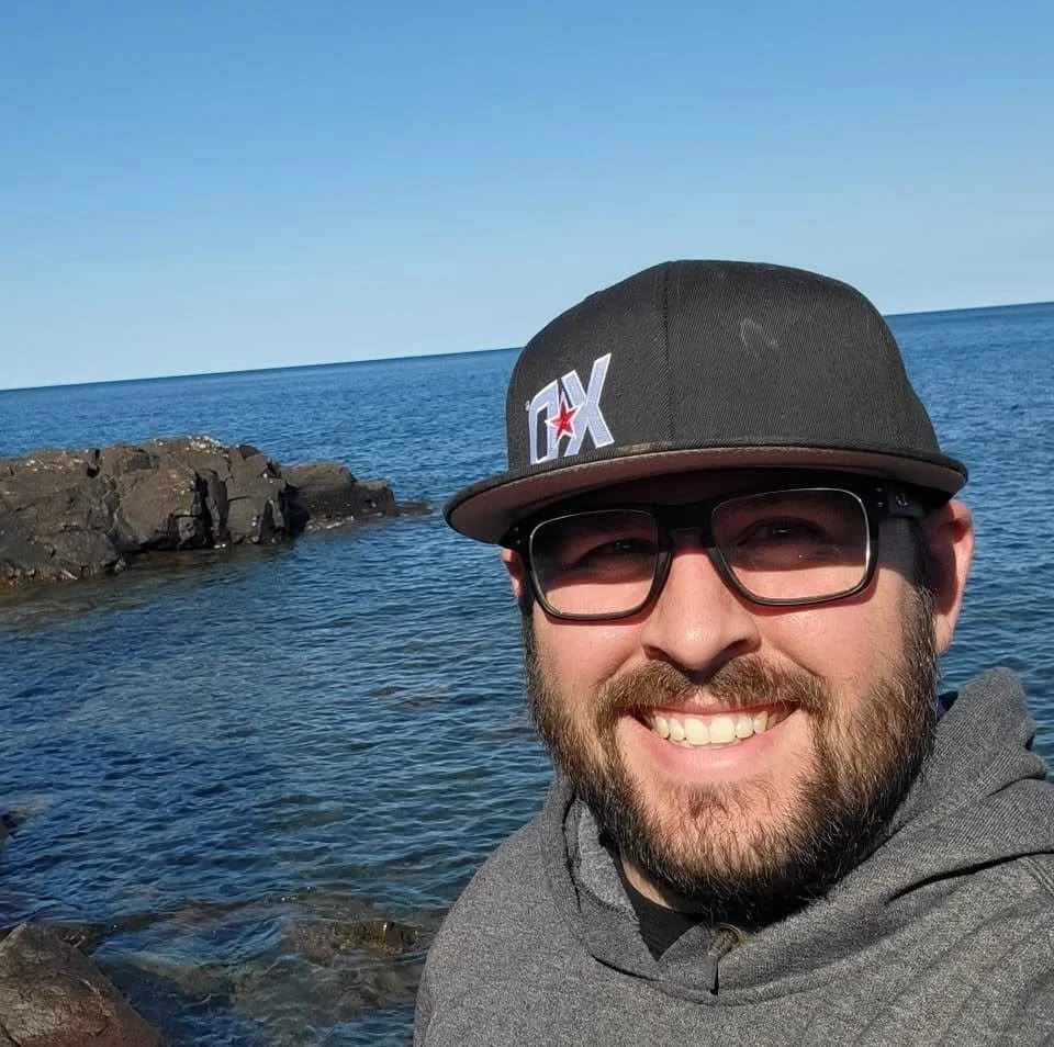 A man smiling at the camera near the ocean with rocks in the background, wearing a black cap with 'DX' on it and sunglasses.