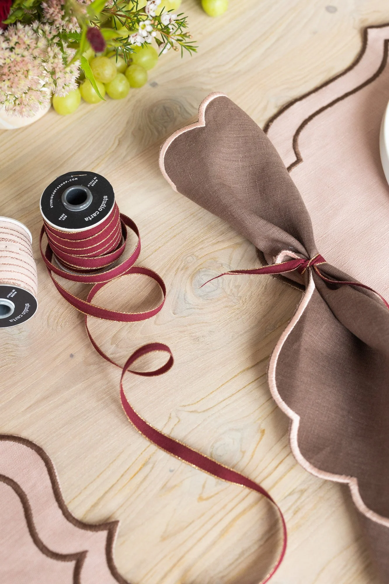 A pink and brown scalloped table runner with a matching napkin tied in a bow, next to spools of pink and red ribbon, on a wooden table with a floral arrangement in the background.