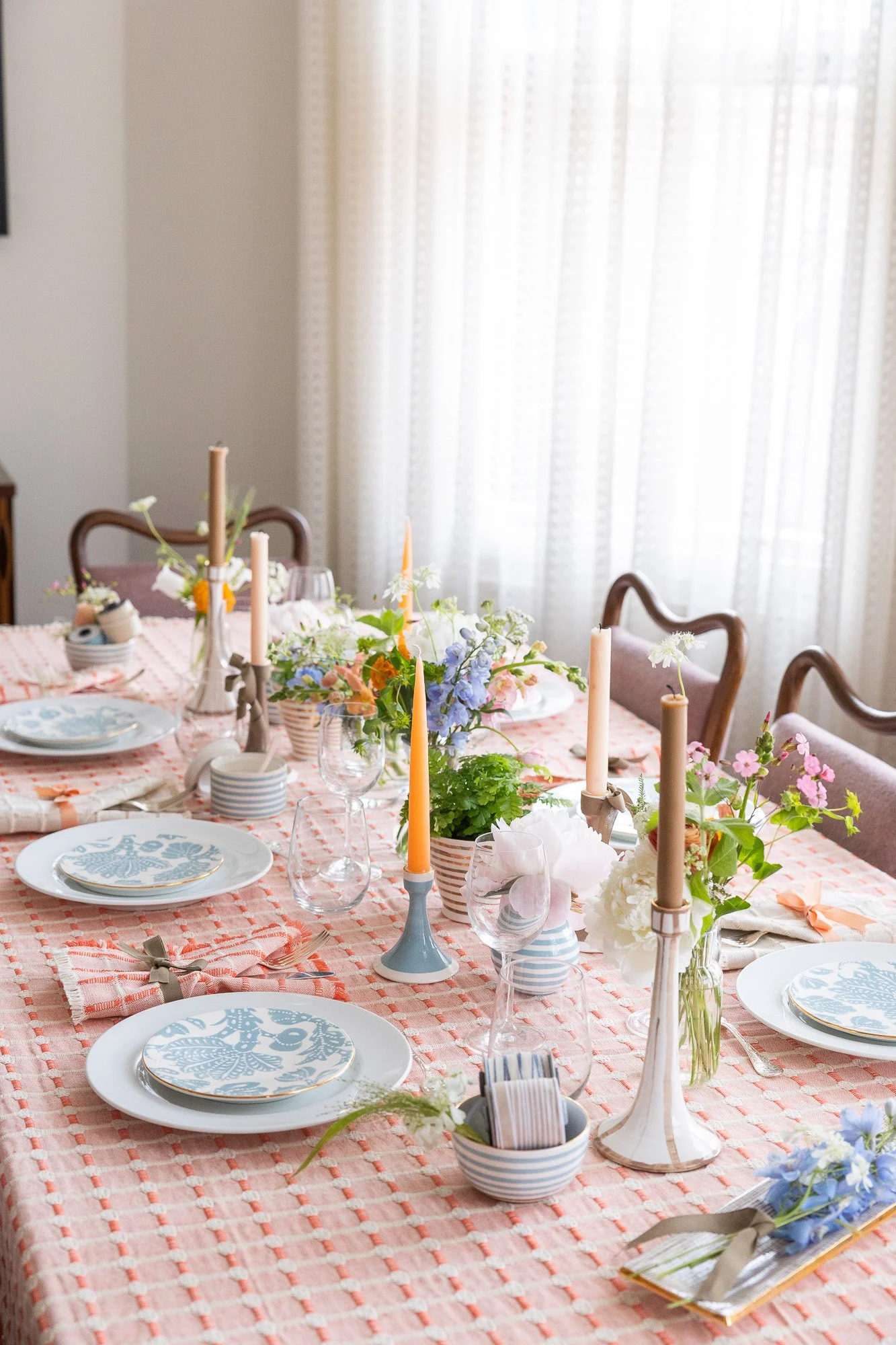 A decorated dining table set for a gathering with pastel-colored candles, floral centerpieces, patterned plates, glassware, and napkins.