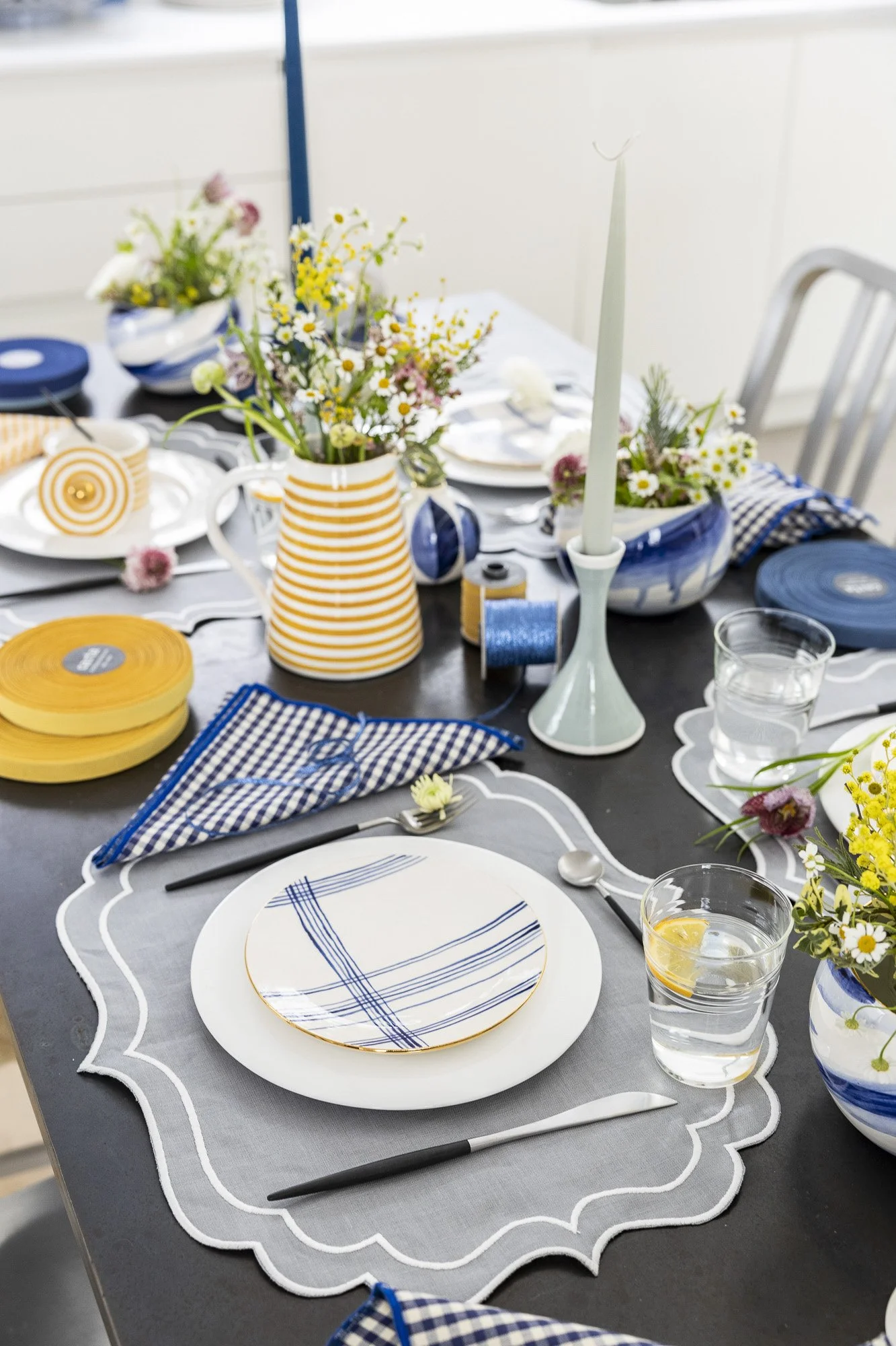 A table set for a meal with plates, glasses of water with lemon slices, blue checkered napkins, and floral centerpieces in blue and yellow vases. There are also stacks of yellow plates, a white candlestick holder with an unlit candle, and various ribbons on the table.