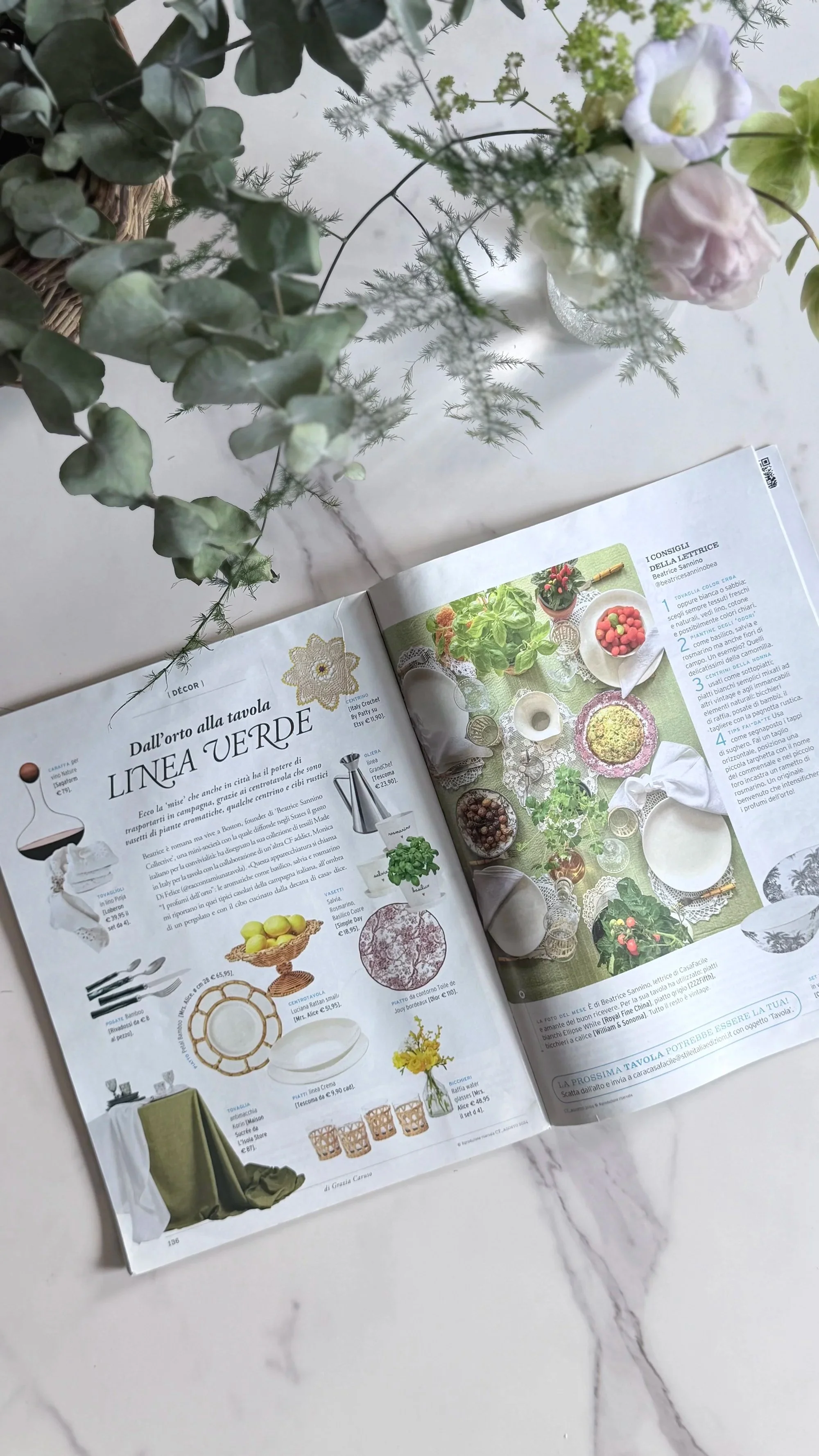 Open magazine showing a table setting with dishes, glasses, and tableware, alongside a plant and floral arrangement on a white marble surface.