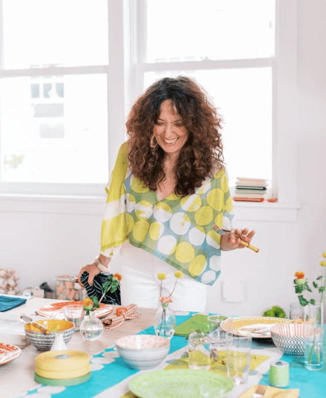 A woman with curly brown hair, wearing a yellow and green polka-dot top, laughing while holding a yellow utensil, standing at a table decorated with colorful dishes and vases.