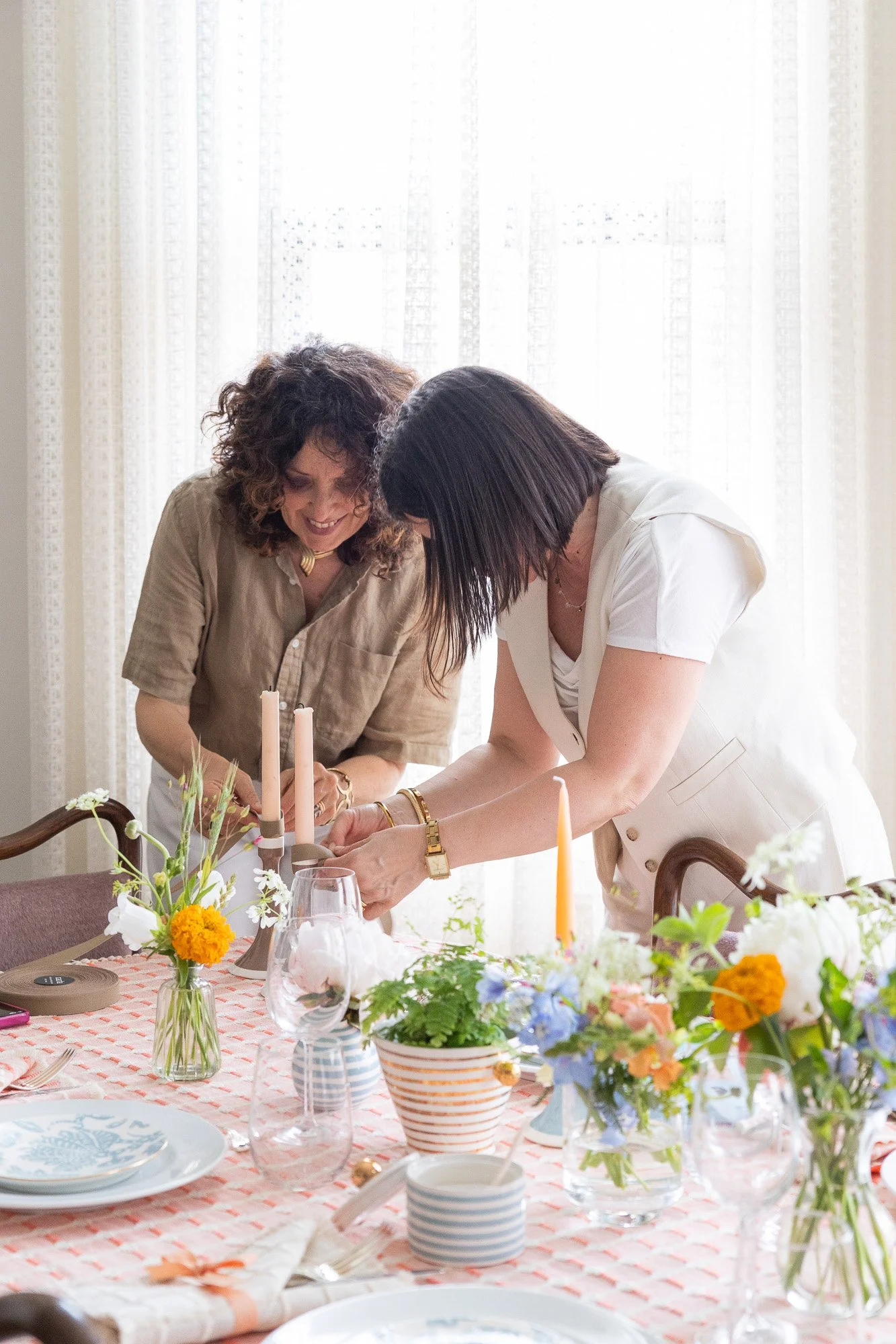 Two women are setting a dining table with plates, glasses, flowers, and candles, smiling and working together near a window with sheer curtains.