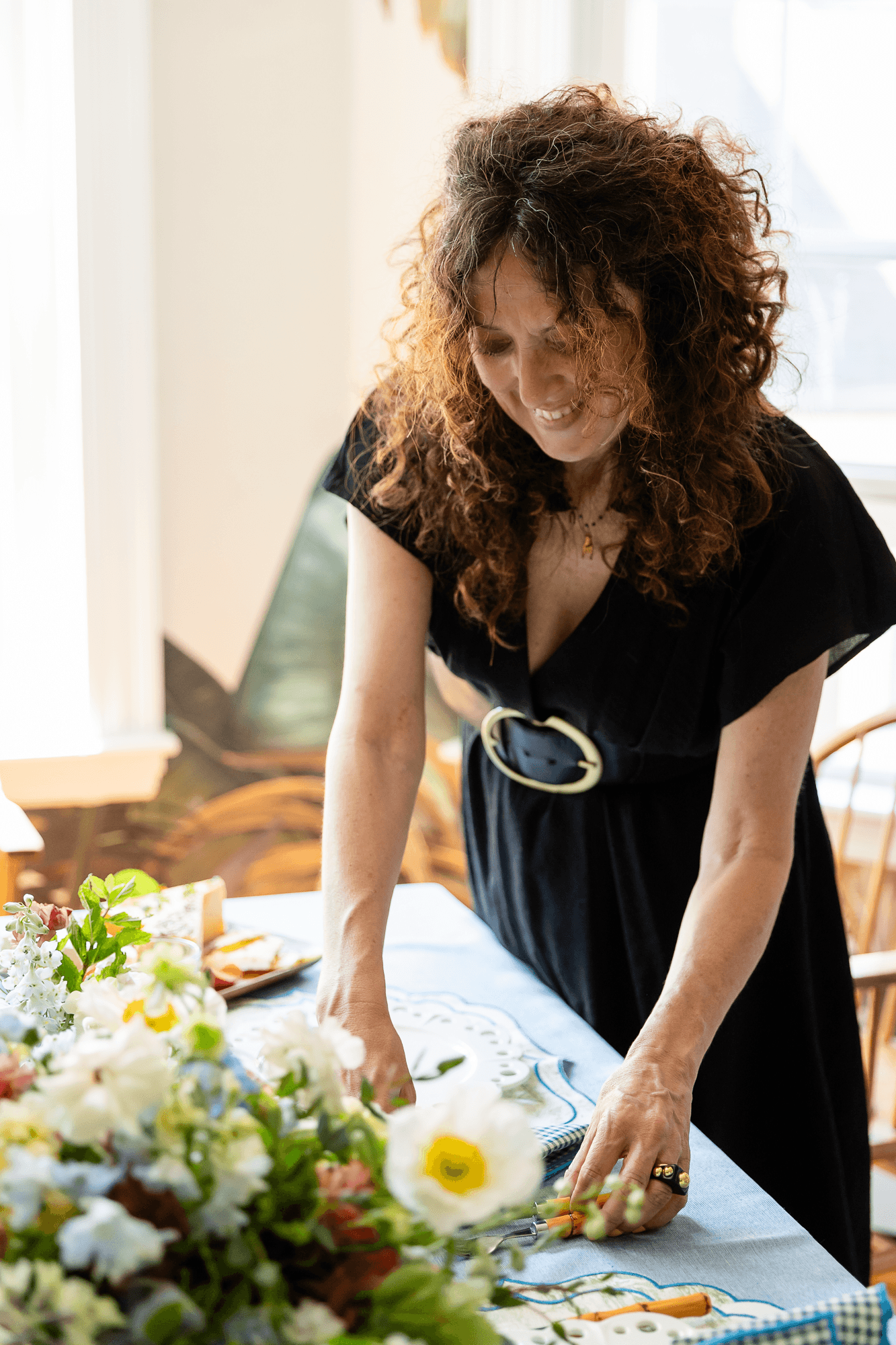 A woman with curly red hair, wearing a black dress with a large belt, is leaning over a table decorated with flowers and arranging items. She appears to be smiling or laughing.