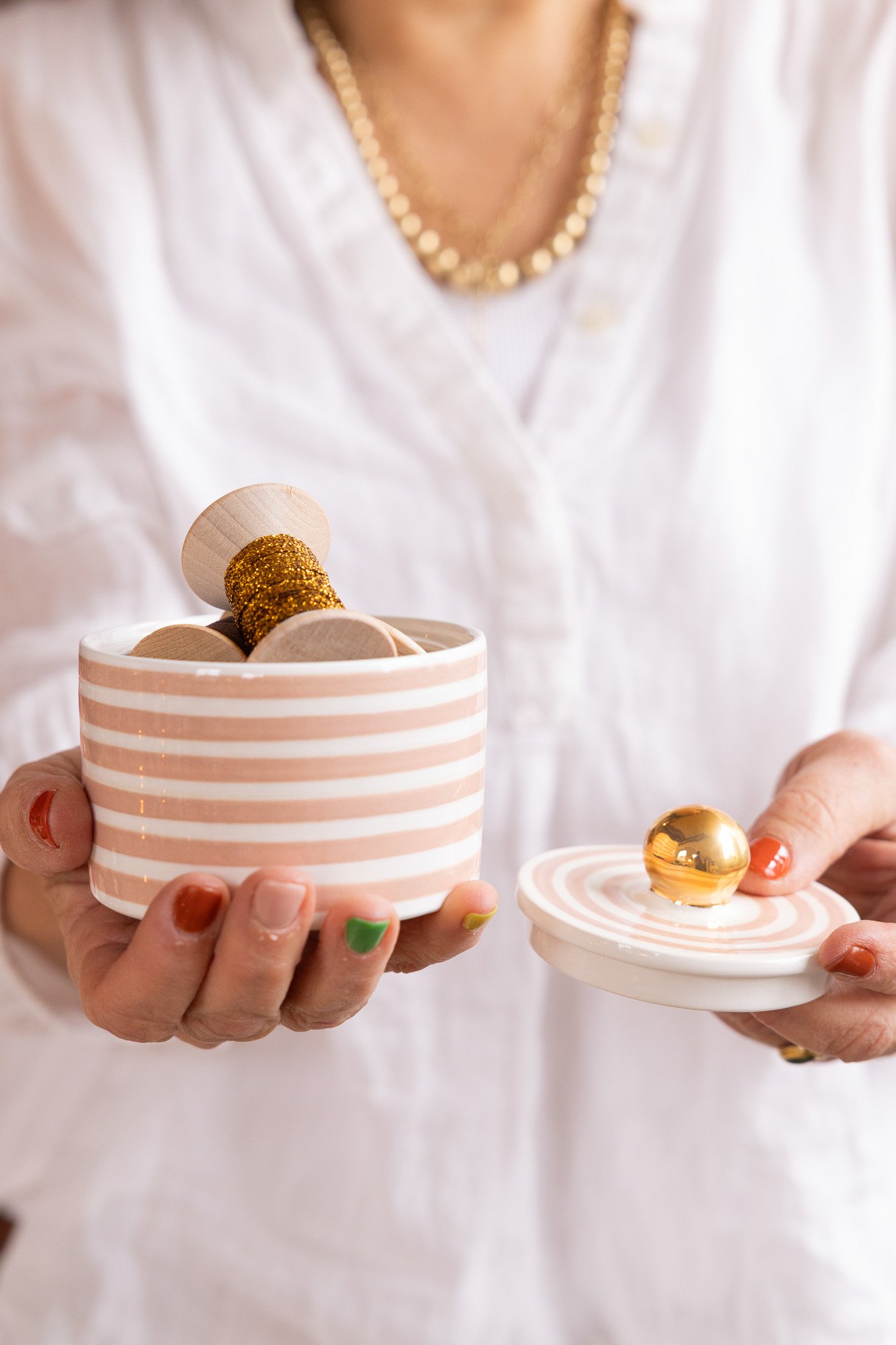 Person holding a pink and white striped jar filled with wooden and glittery gold ornaments, with a matching decorated lid in the other hand.