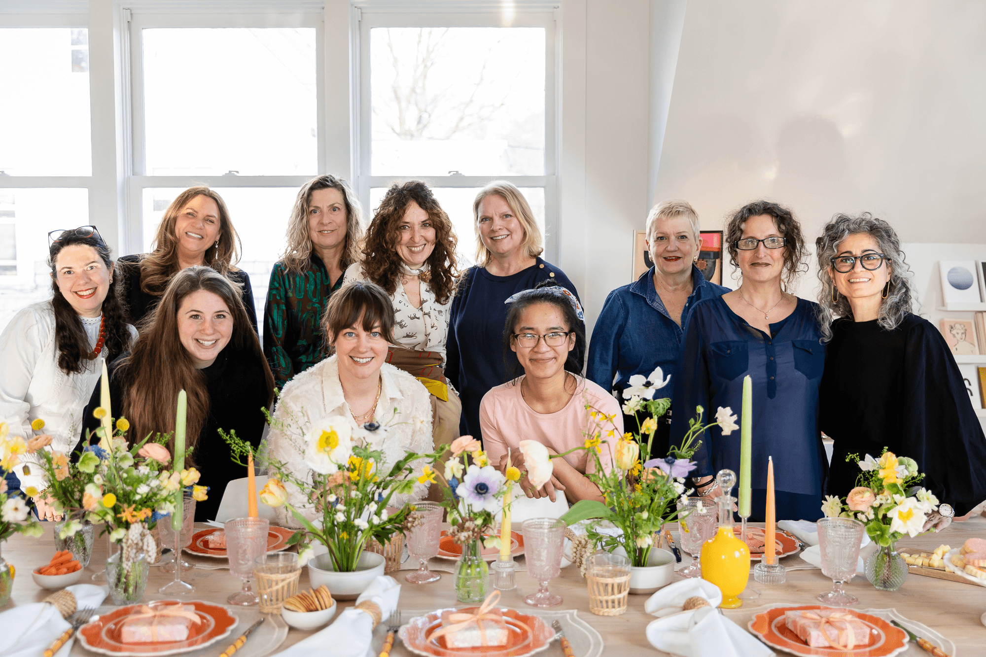 Group of women gathered around a decorated table with flowers, candles, and tableware in a bright room.