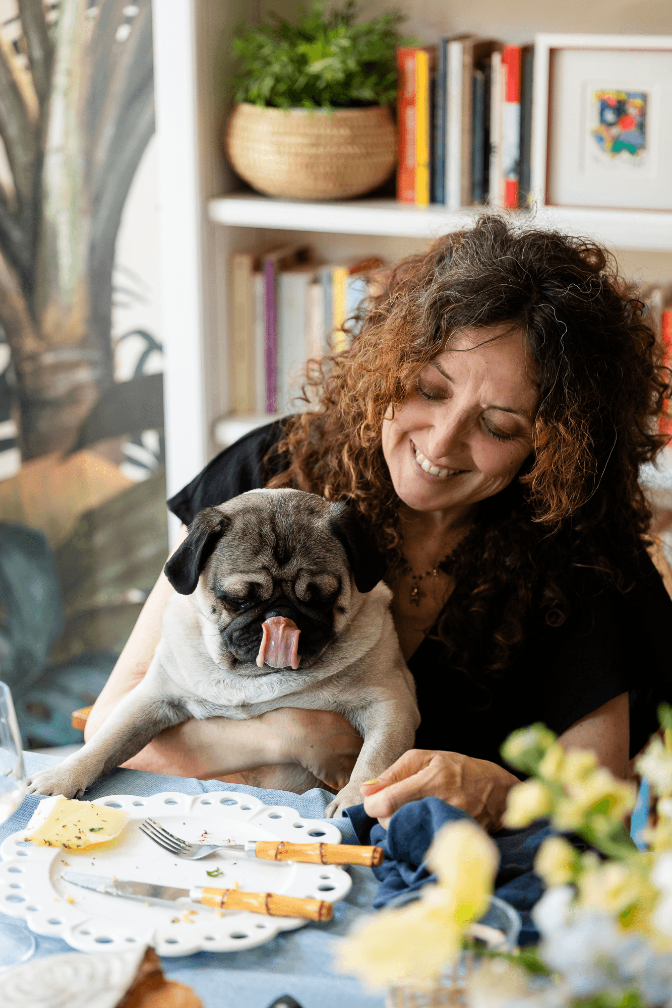 A woman smiling and holding a pug at a table with a partially eaten piece of cheese, a fork, and a knife. The setting appears to be a cozy room with a bookshelf and a large plant in the background.