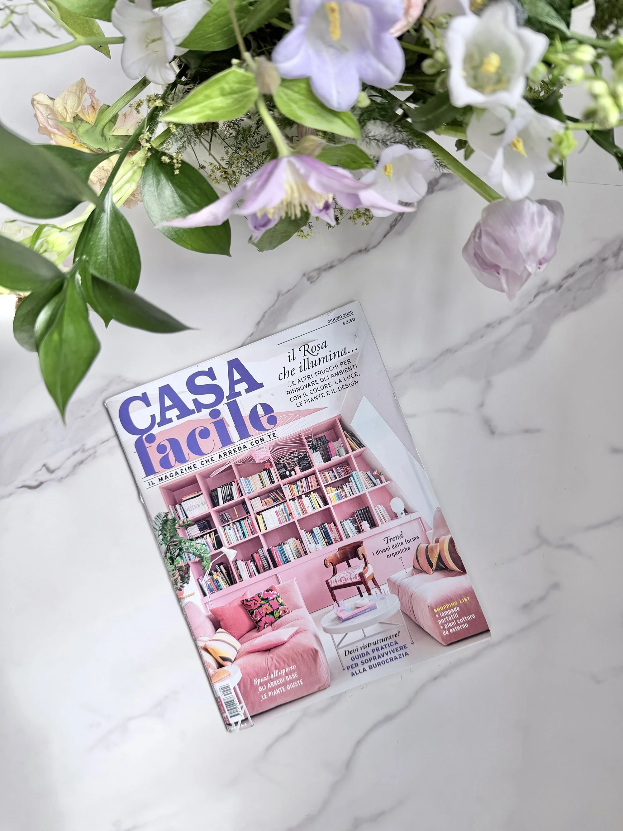 A magazine titled 'Casa Facile' on a white marble surface, partially covered by a bouquet of flowers with pink, white, and purple blossoms and green leaves.