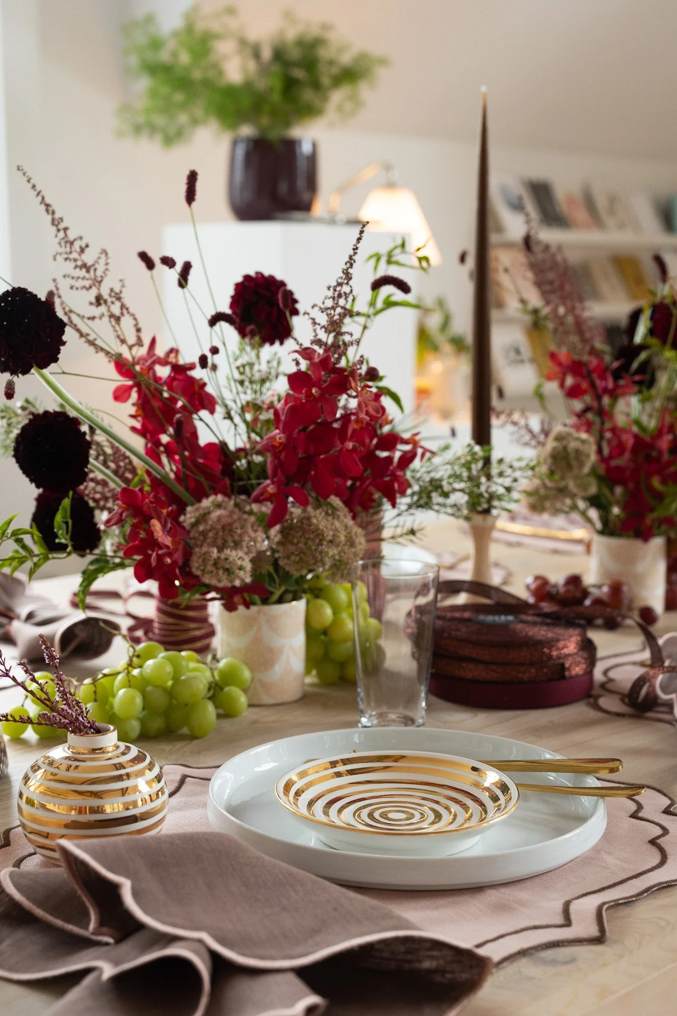 A decorated dining table with floral arrangements, a white plate with a gold spiral pattern, a gold spoon, a glass of water, and a small vase with gold stripes.