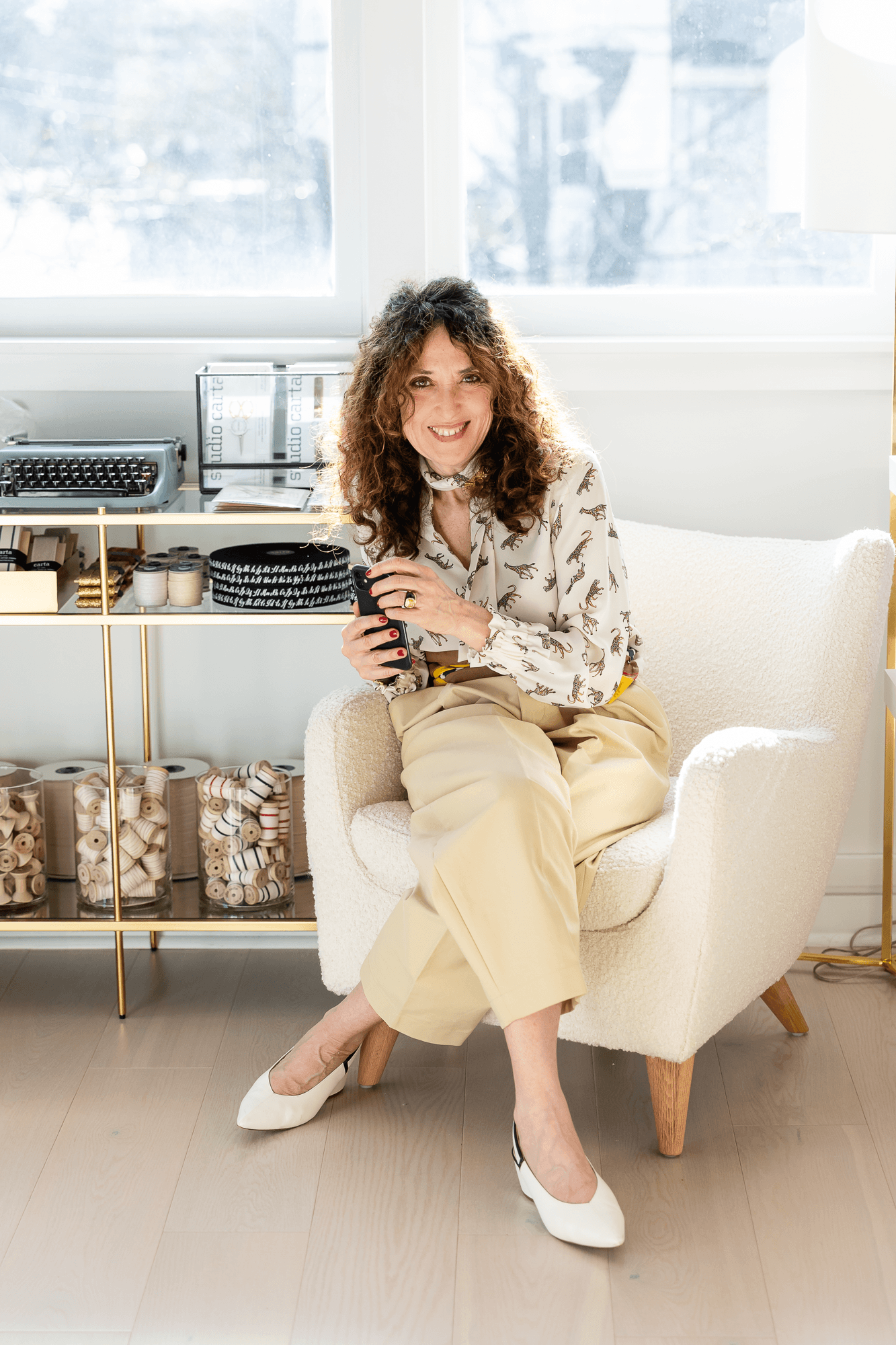 A woman with curly hair sitting on a cream-colored armchair, holding a smartphone, smiling, in a bright room with large windows and a shelving unit with stationery items.