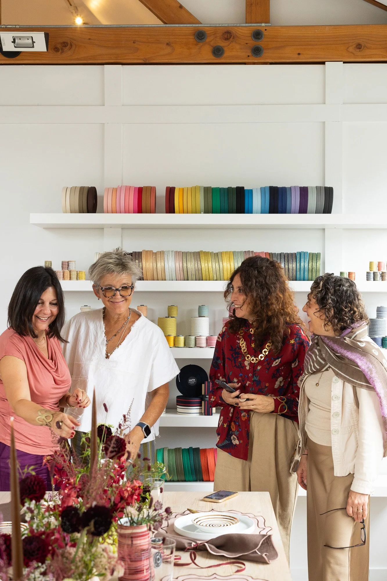 Four women in a craft store, with shelves of colorful ribbons behind them, are looking at a floral arrangement on a table.