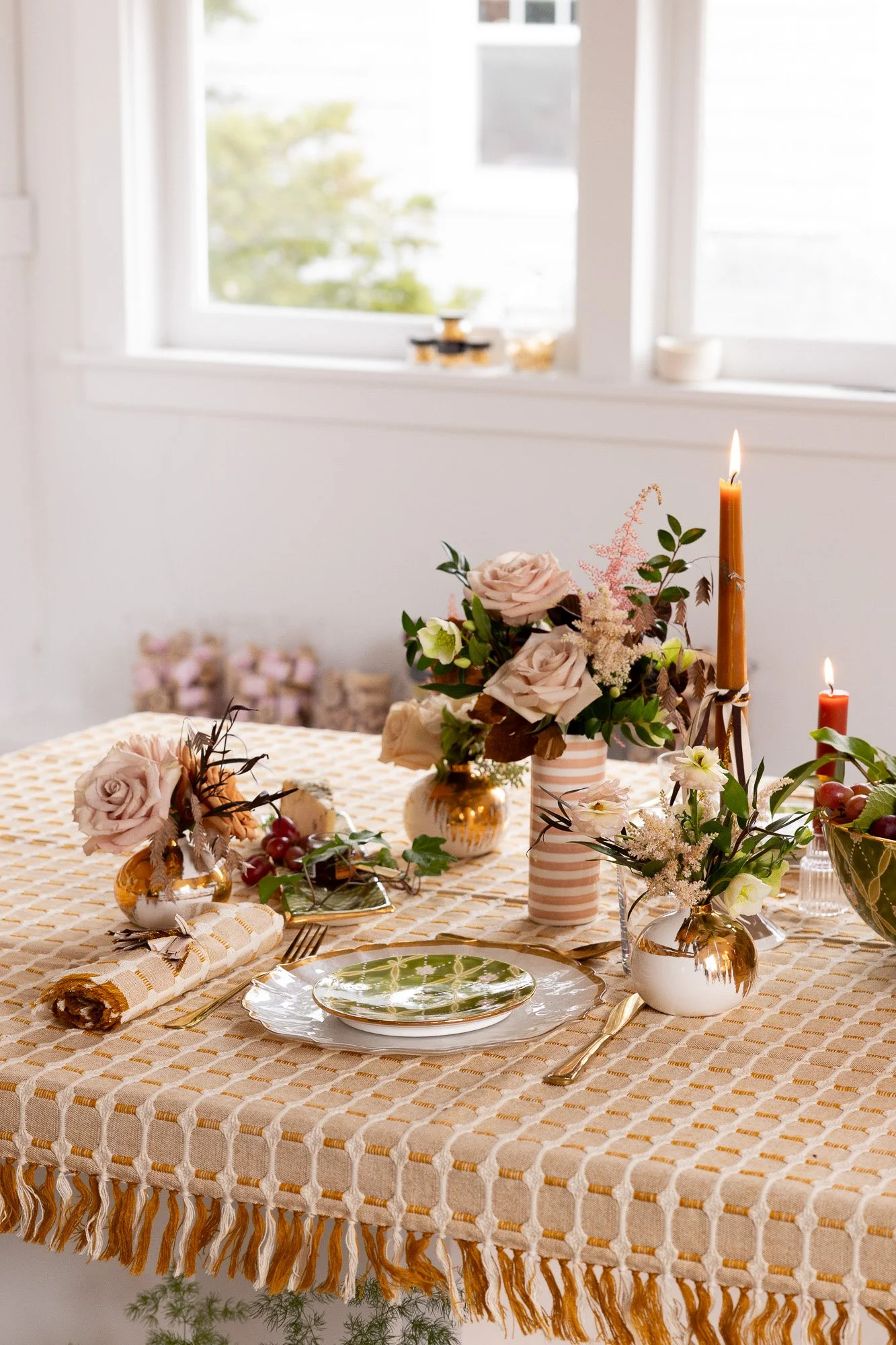 A decorated dining table with gold and pink vases holding pink and white flowers, gold cutlery, a rolled napkin, a green and white patterned plate, lit candles, and a fruit bowl in a bright room with large windows.