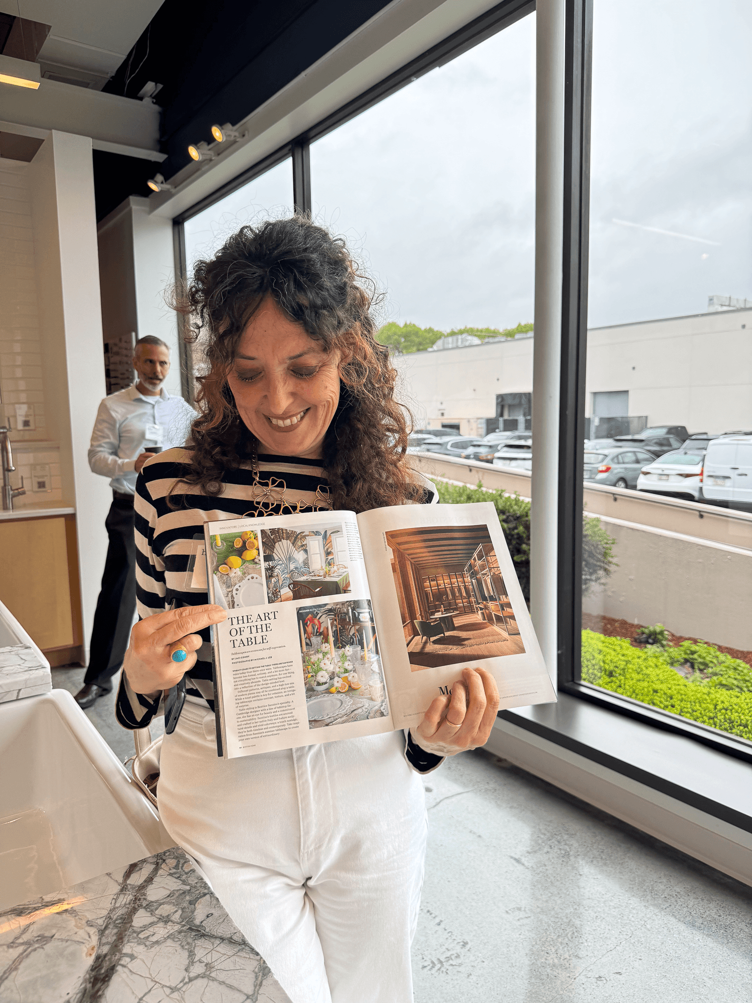 Woman with curly hair smiling as she looks at a magazine about interior design, with a man in the background near large windows in a modern, well-lit room.