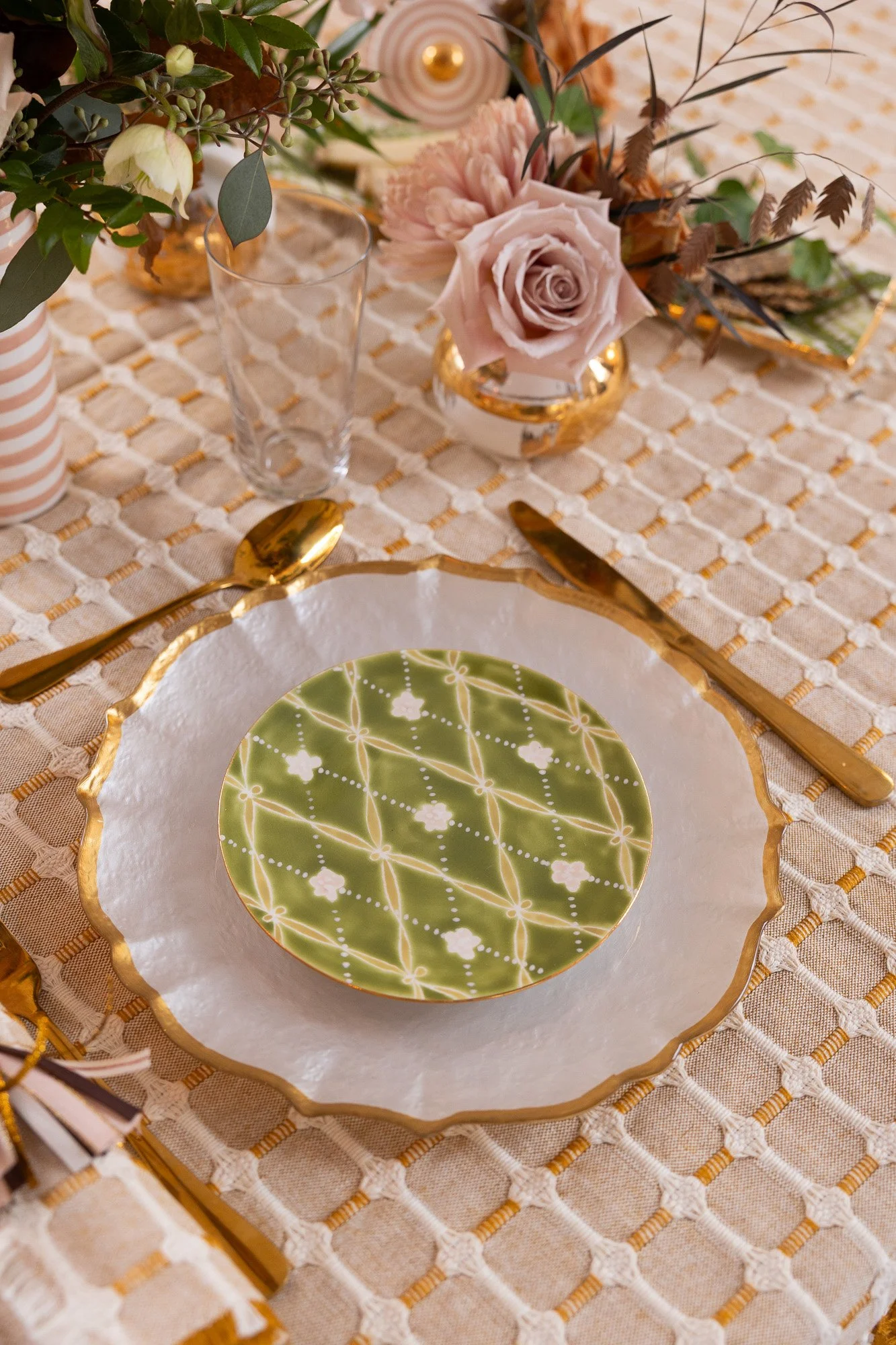 Elegant table setting with a decorative green and gold plate, gold utensils, a floral centerpiece with pastel roses, and a beige tablecloth.