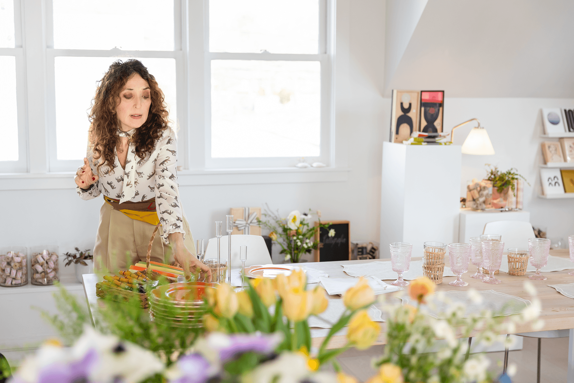 Woman preparing a table with glassware and plates in a well-lit, decorated room with flowers and art on the walls.