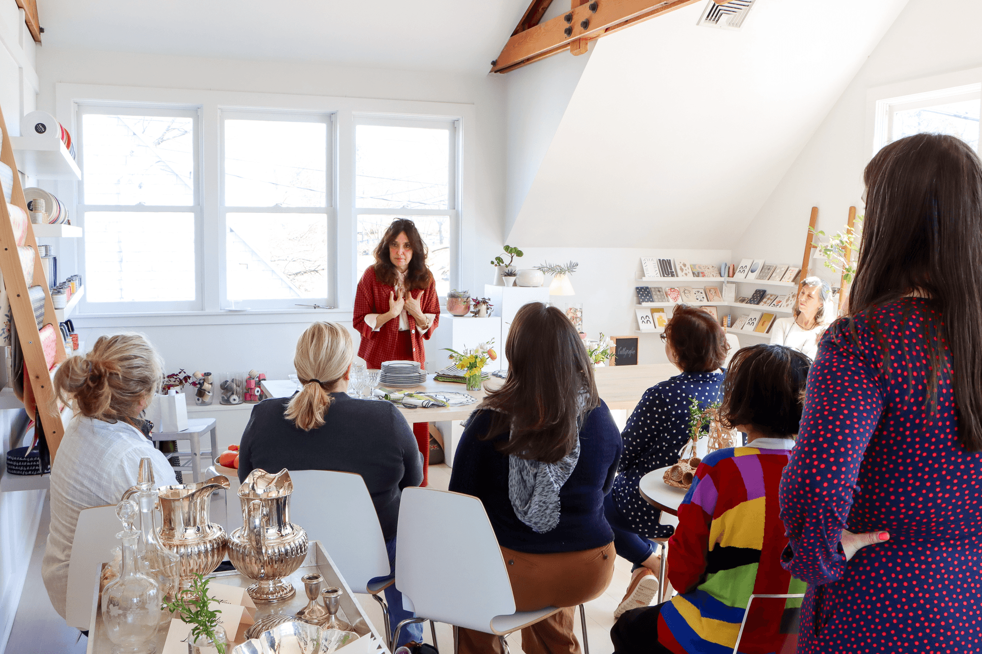 A woman giving a presentation to a group of women seated around a table in a bright room with large windows, white walls, and shelves with books and decor.