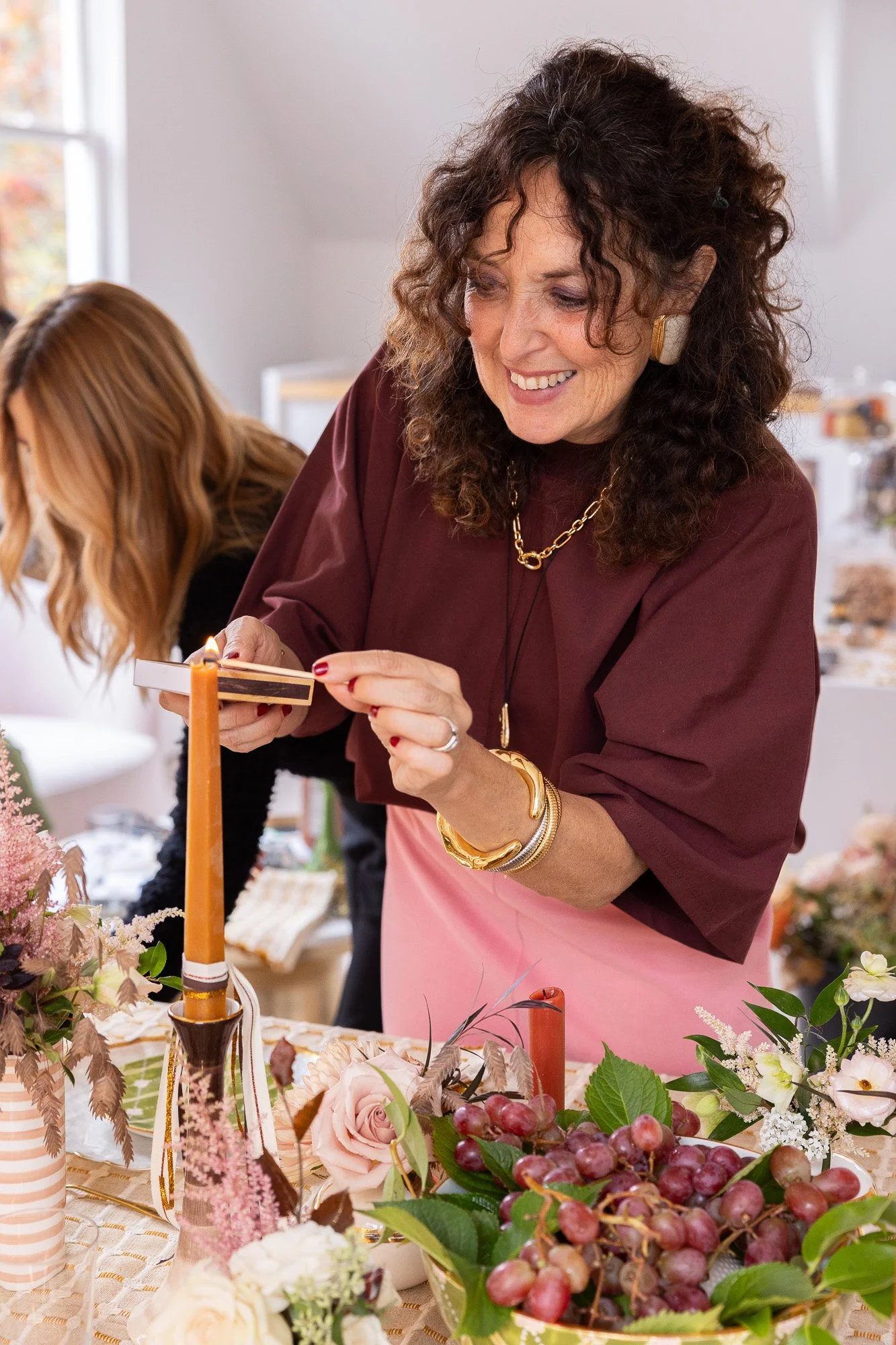 A woman in a dark red top and pink apron lighting a tall candle on a table decorated with flowers, grapes, and candles, while another person with long blonde hair works in the background.