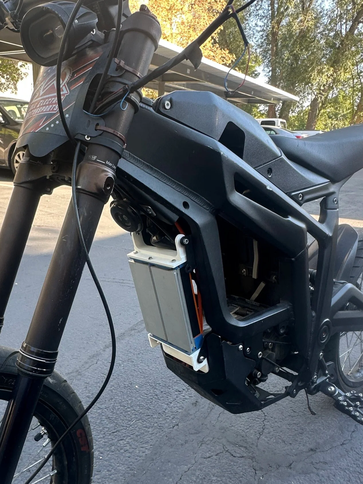 An electric motorcycle with the side panel removed revealing the battery inside, parked outside on a paved surface in a lot with trees and cars visible in the background.