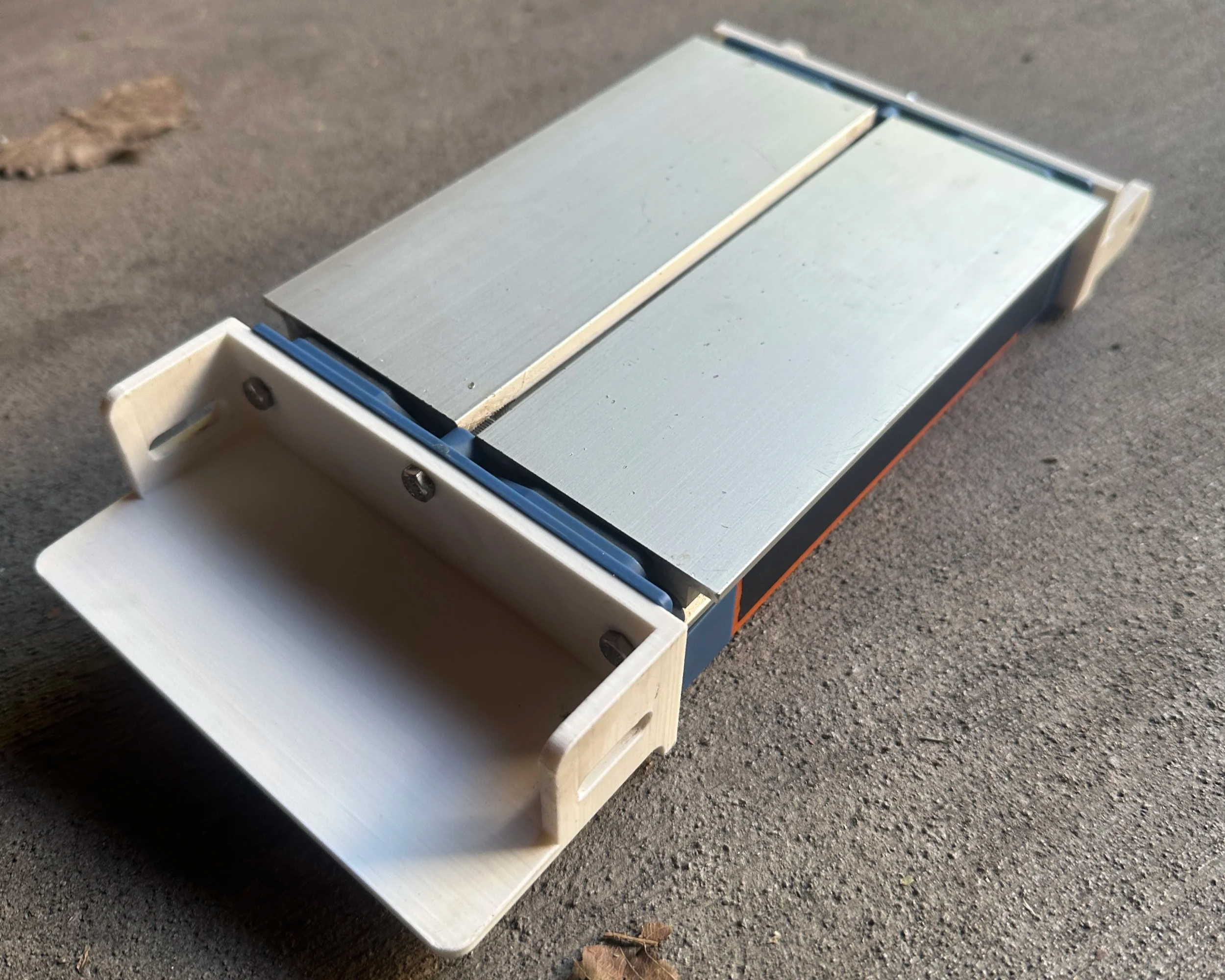 A close-up of a small, rectangular, wooden drawer slide mechanism with metal rails, placed on a rough outdoor surface.