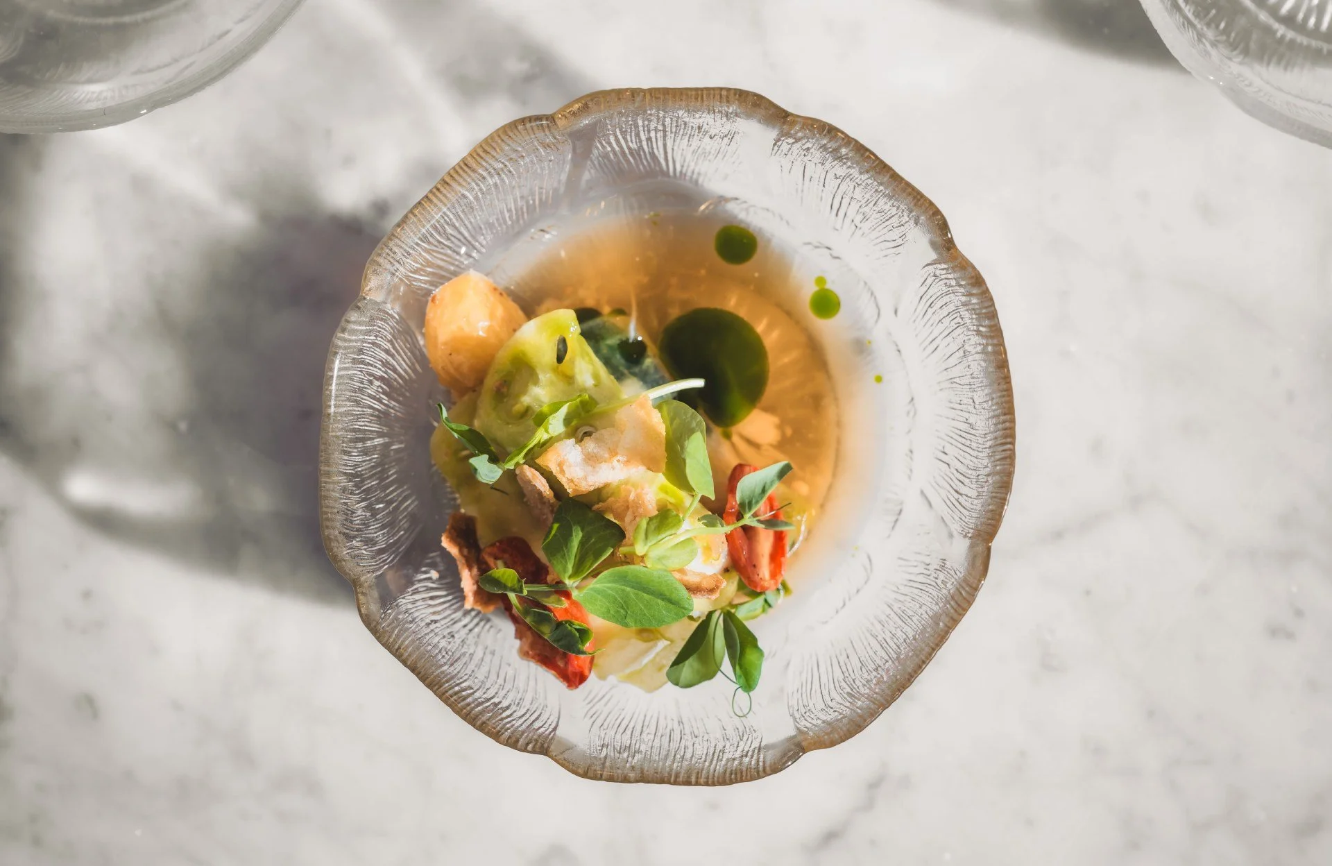 A glass bowl with scalloped edges containing a gourmet dish with various vegetables, greens, and a light sauce, on a white marble surface.