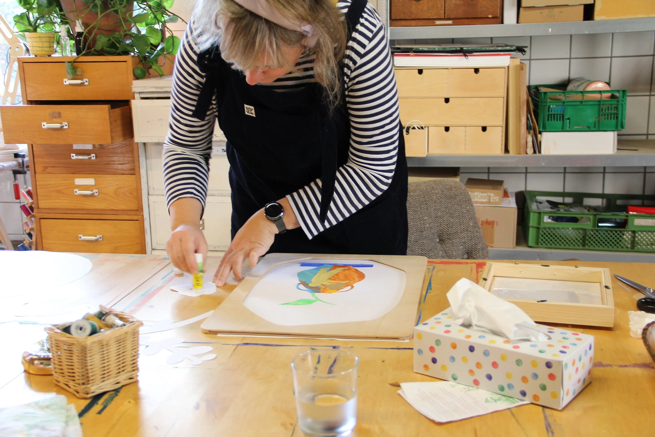 A woman working on an art project at a wooden table, surrounded by art supplies, tissue box, glass of water, and a colorful drawing of a bird.