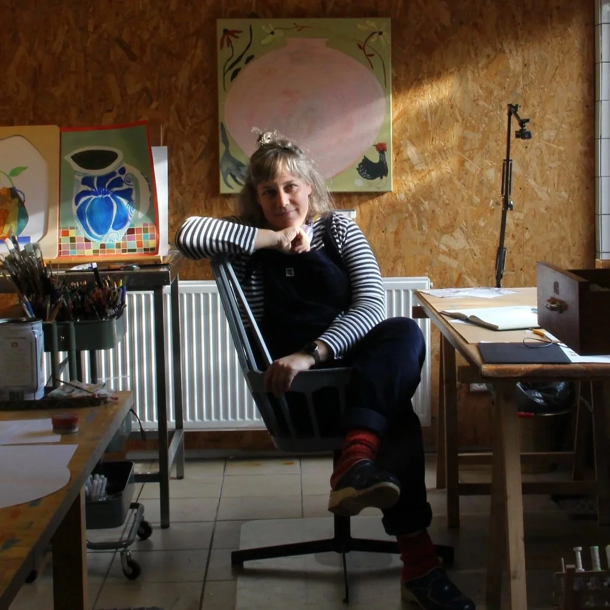 A woman with gray hair wearing navy blue dungarees and striped shirt is sitting on an office chair in an art studio, leaning her head on her hand and smiling. There are colorful artwork and art supplies on the tables and shelves around her.