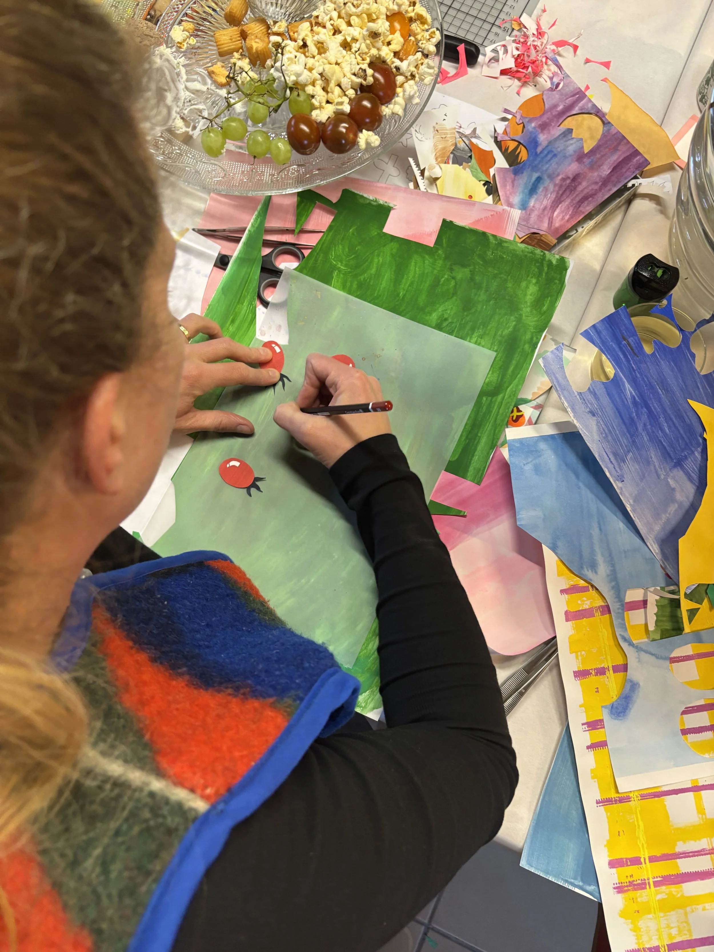 A person painting cartoon rose hips on a green background at a craft table filled with art supplies and decorative paper.