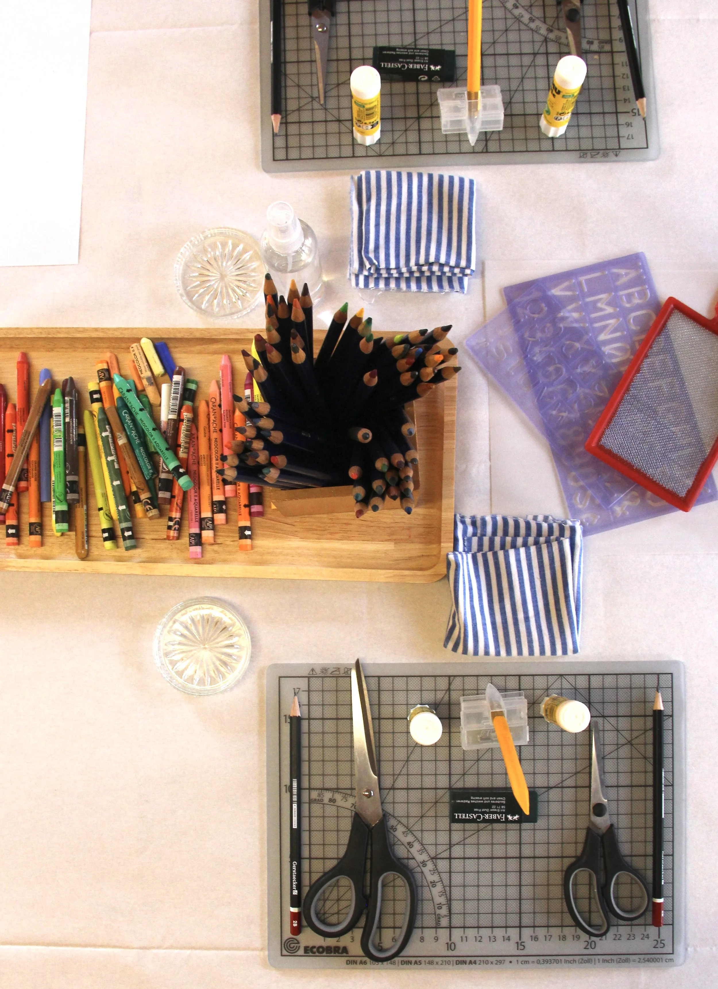 Top view of a craft workspace with markers, colored pencils, scissors, glue, and stencils on a table with a cutting mat. A table set up for a workshop.