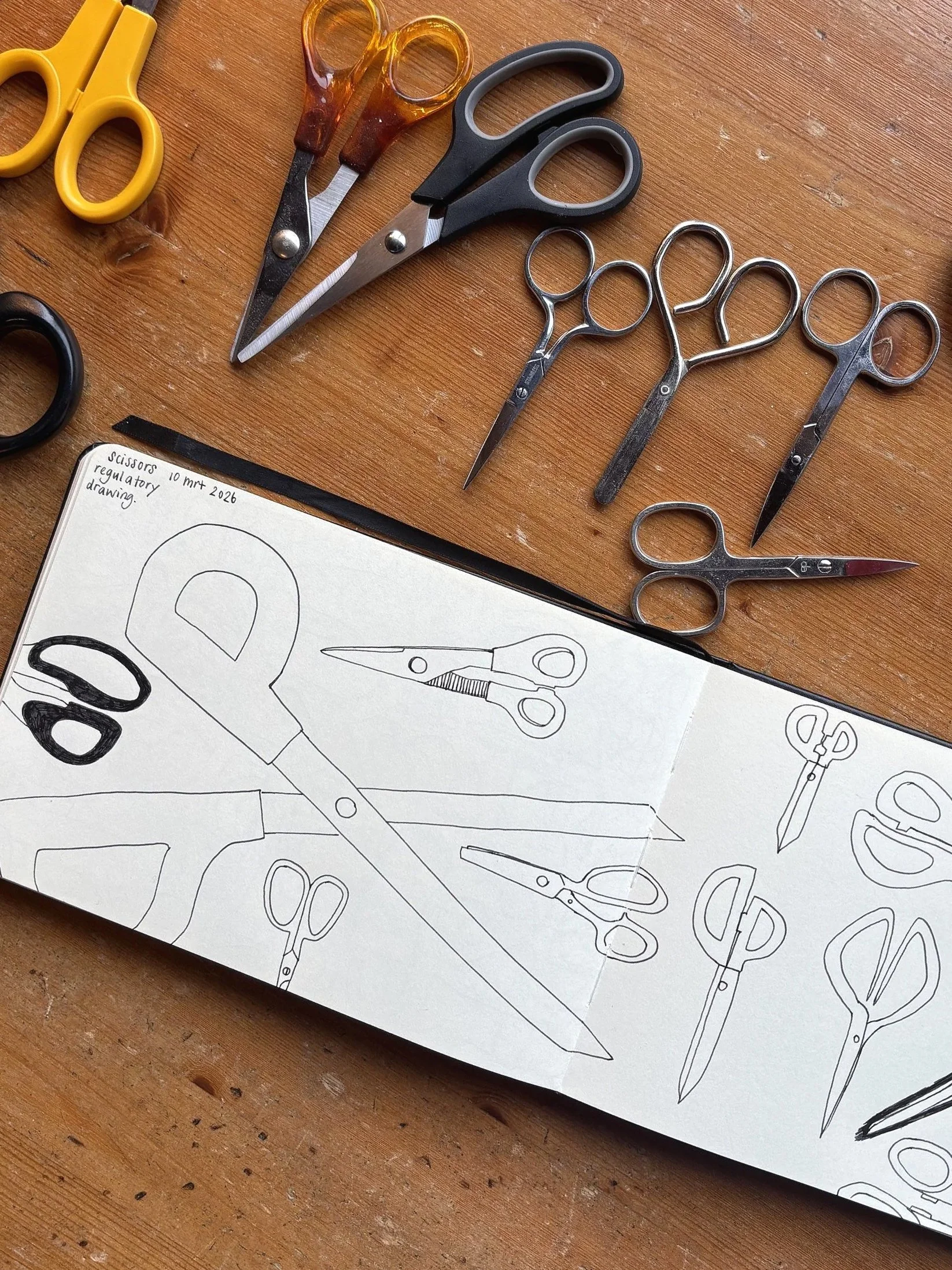 A sketchbook showing various drawings of scissors, with real scissors of different sizes and colors placed above on a wooden surface.