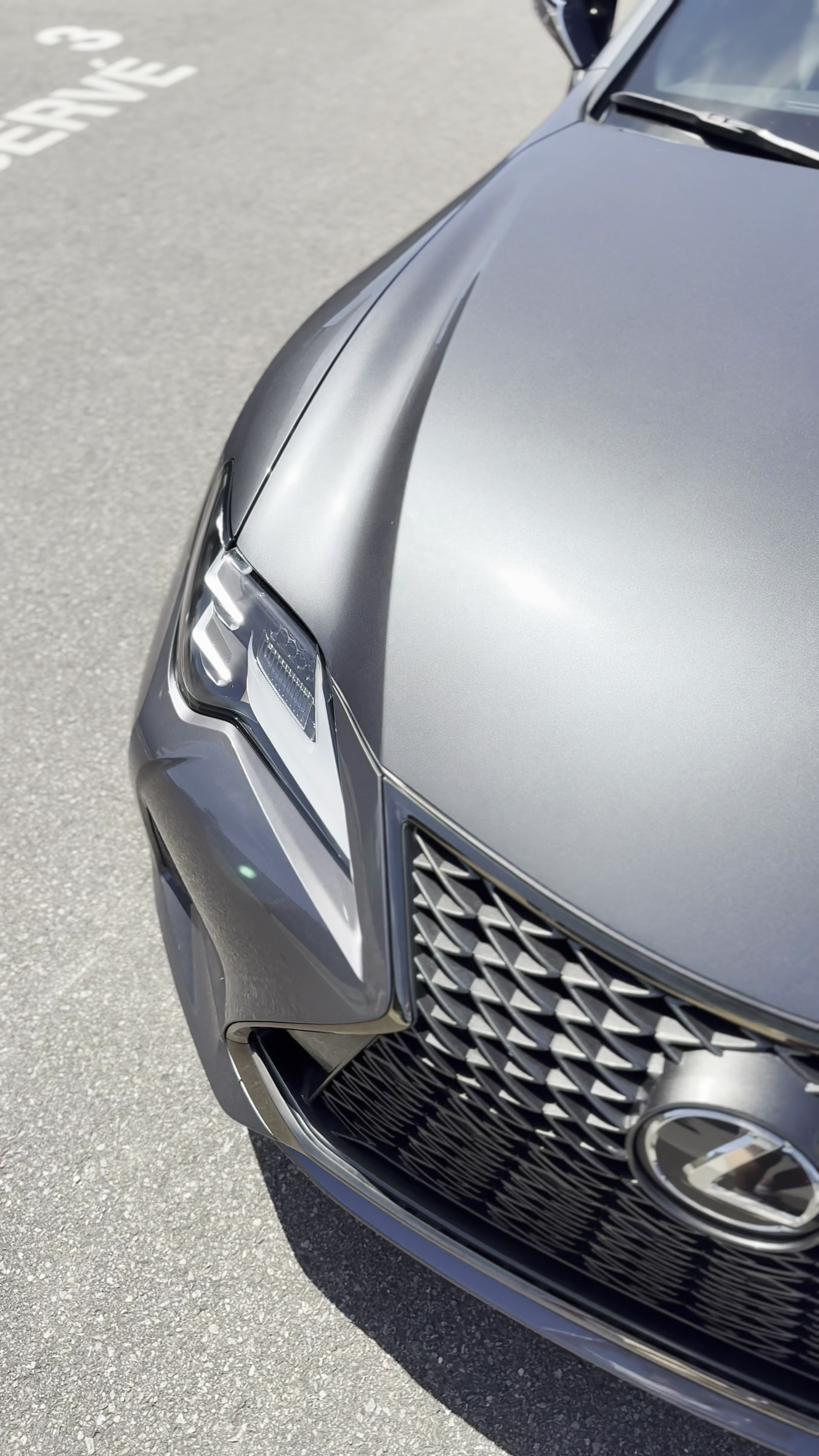 Une voiture grise de luxe, vue partielle de l'avant, stationnée sur une surface en asphalte, avec une focus sur la calandre, le phare et le capot.