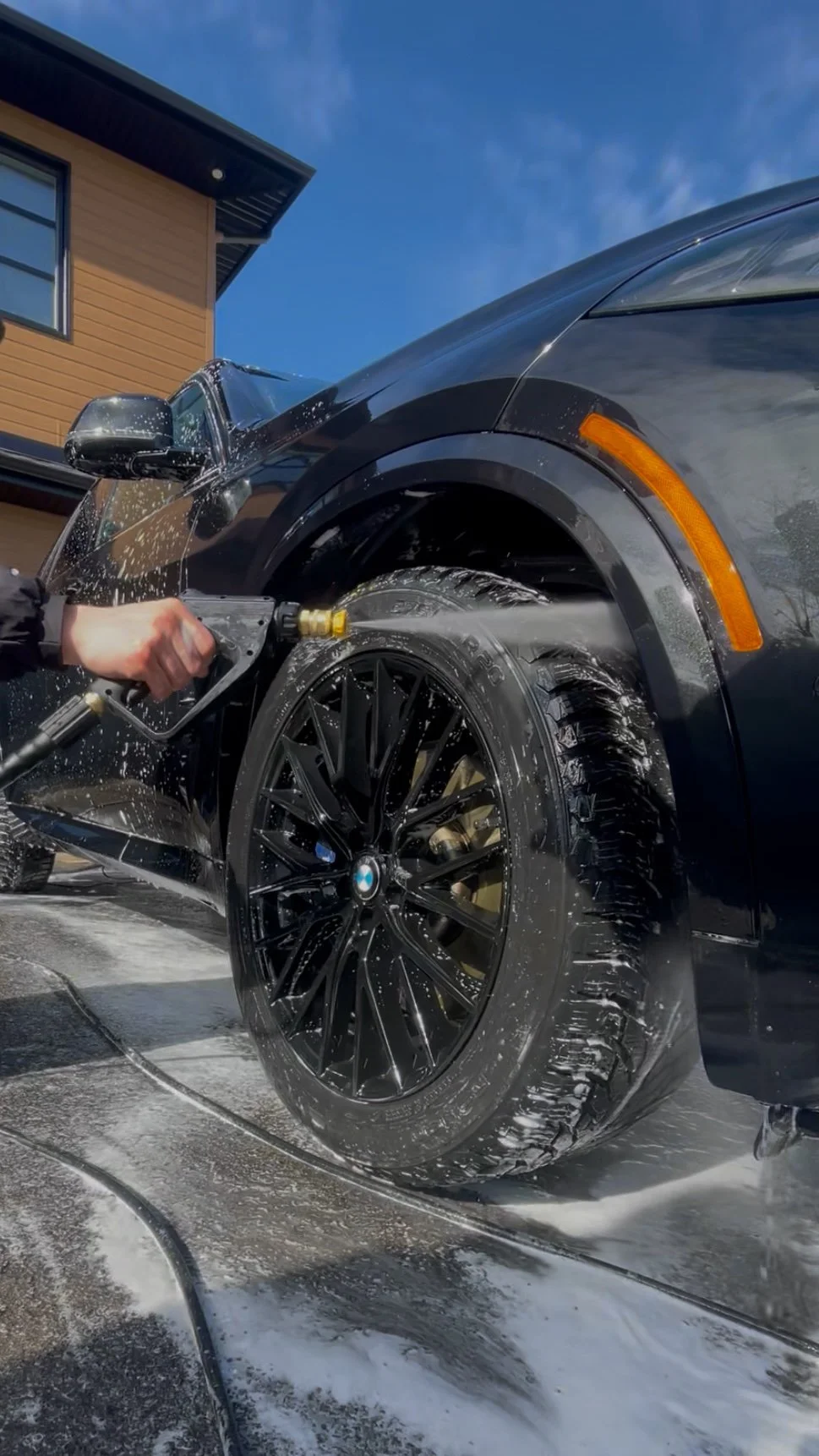 Une personne lave une voiture noire avec un jet d'eau à haute pression dans une maison en arrière-plan sous un ciel bleu.