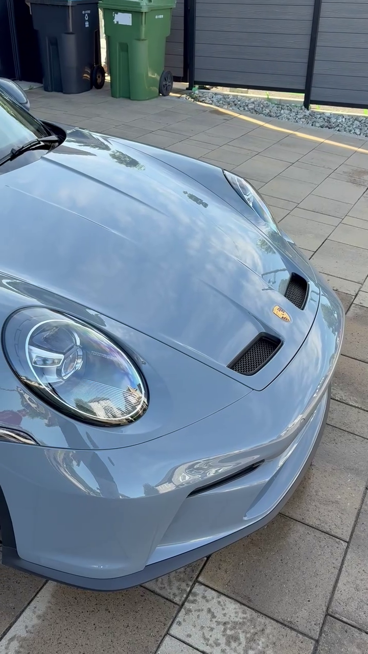 Voiture grise de sport Porsche avec surfaces réfléchissantes, stationnée sur une allée pavée devant une clôture en métal et des poubelles vertes et noires.