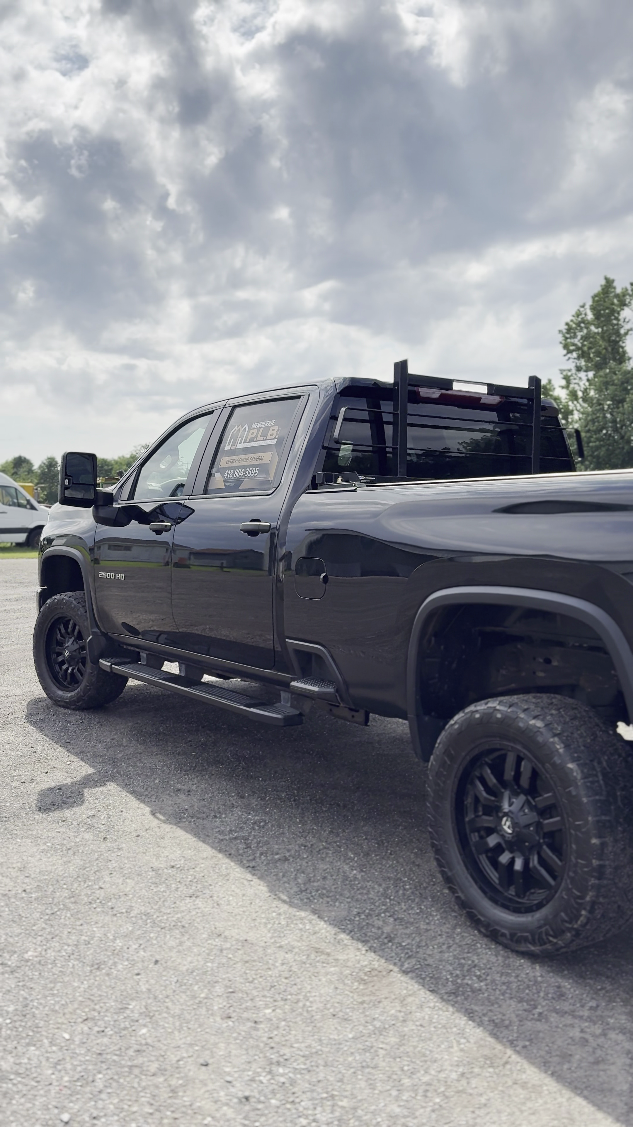 Photo d'un camion noir de taille moyenne avec des pneus tout-terrain, stationné sur une surface asphaltée. Le ciel est nuageux et il y a quelques véhicules en arrière-plan.